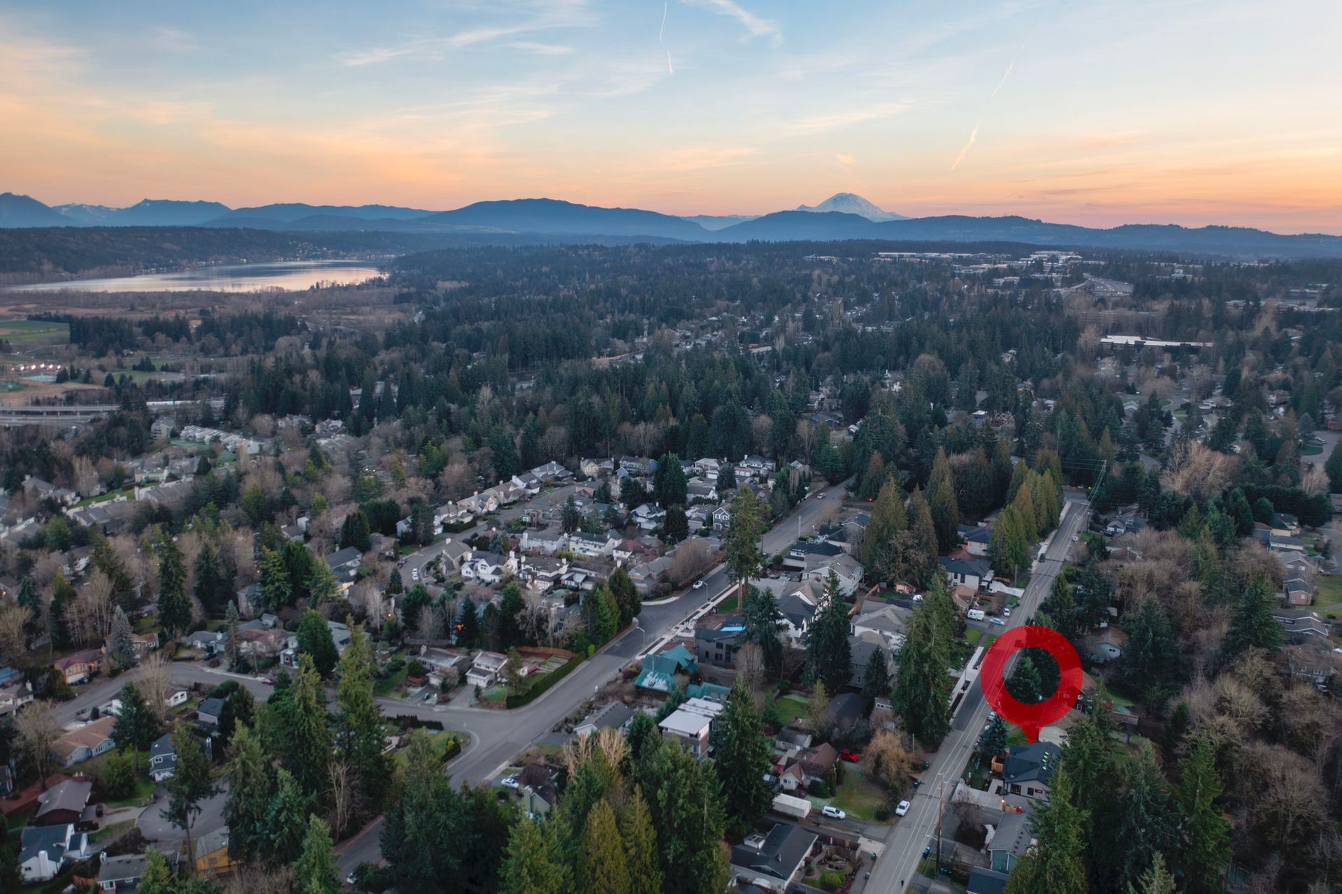 Aerial view of a residential neighborhood with houses and trees. Mountains and a lake are visible in the distance.