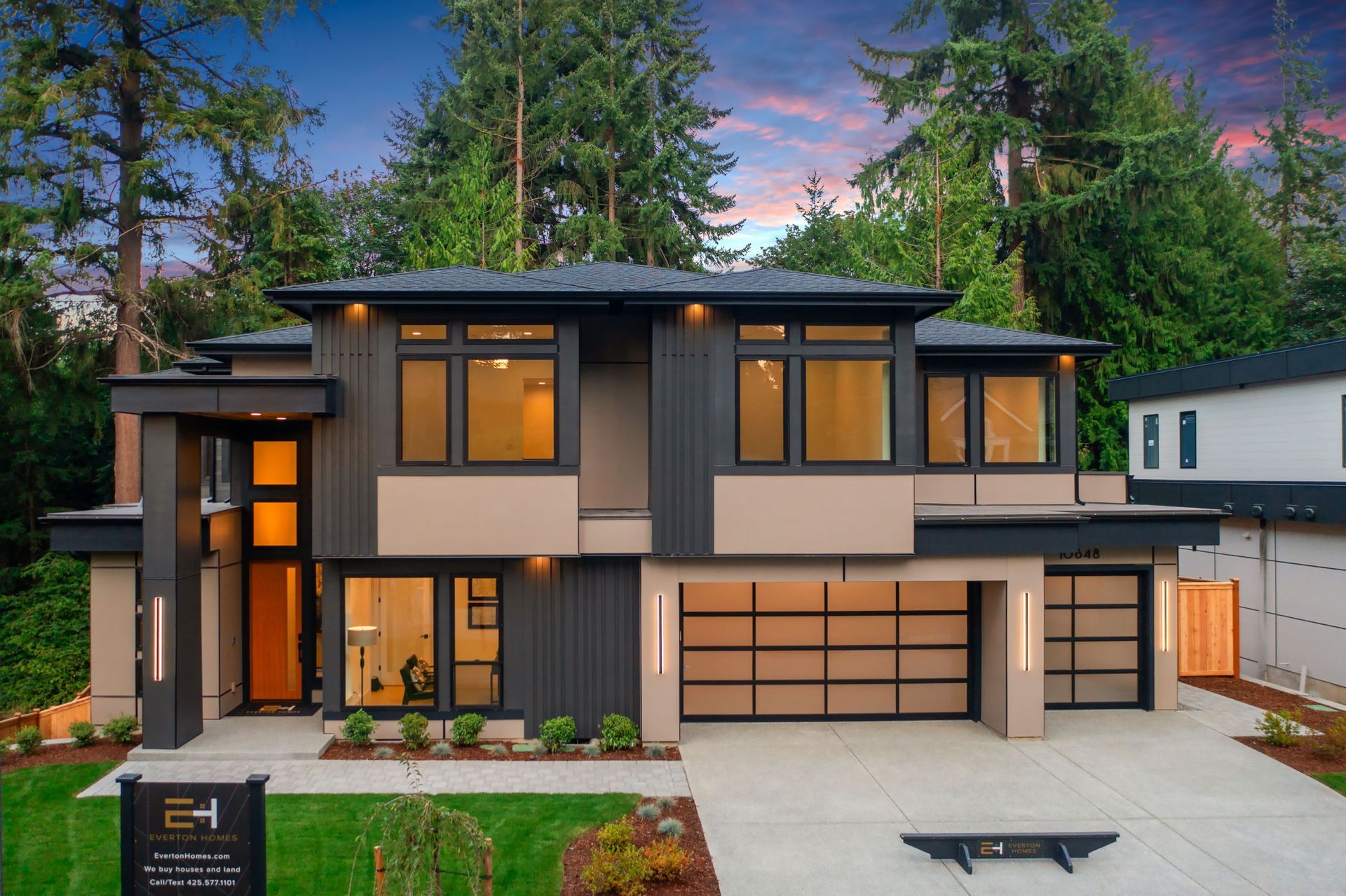 Modern two-story house with gray and beige exterior, large windows, garage, and lush green surroundings.