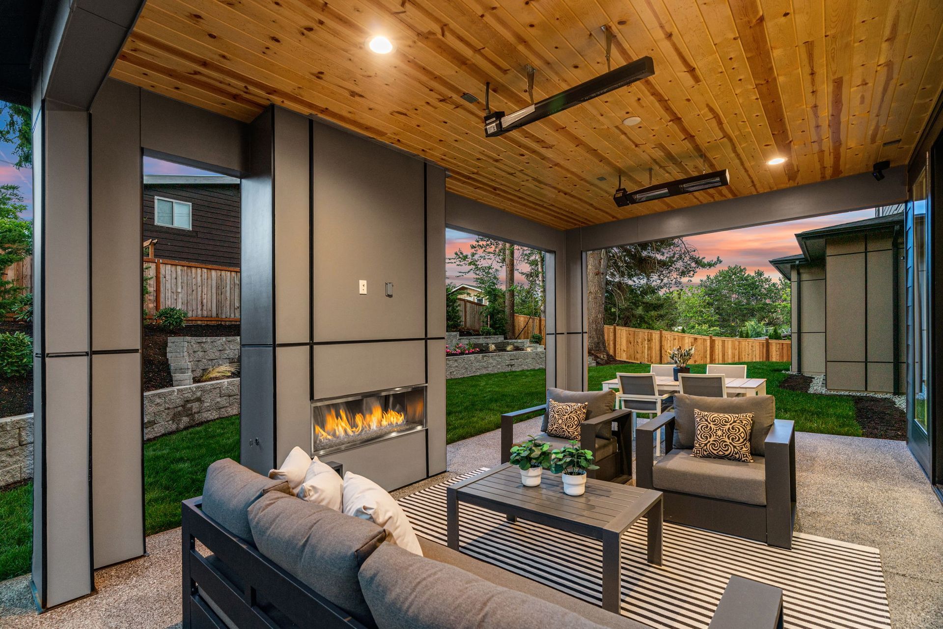 Modern covered patio with fireplace, seating area, wood سقف, and backyard garden at sunset