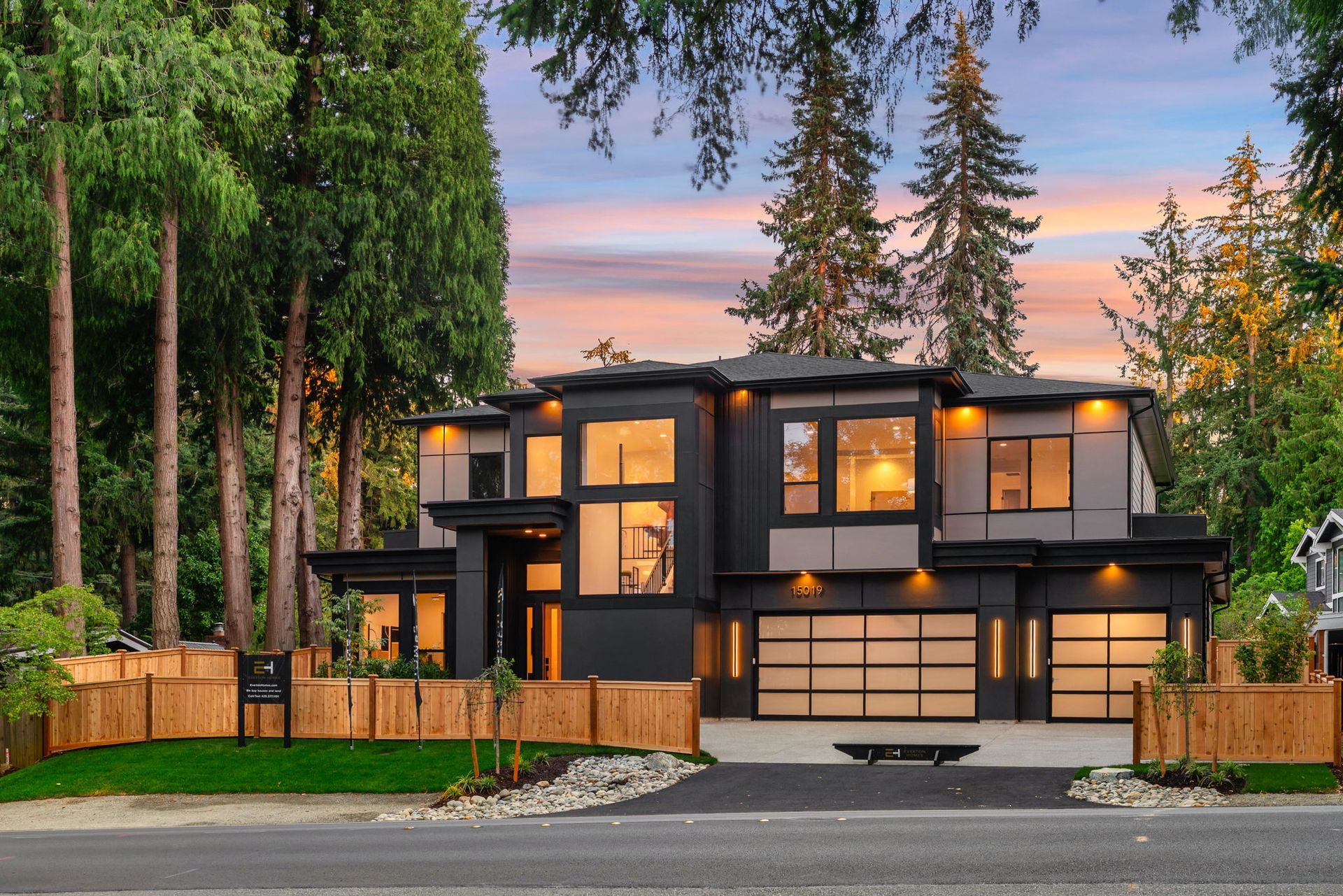 Modern two-story house with warm lights, dark trim, and tall trees at sunset
