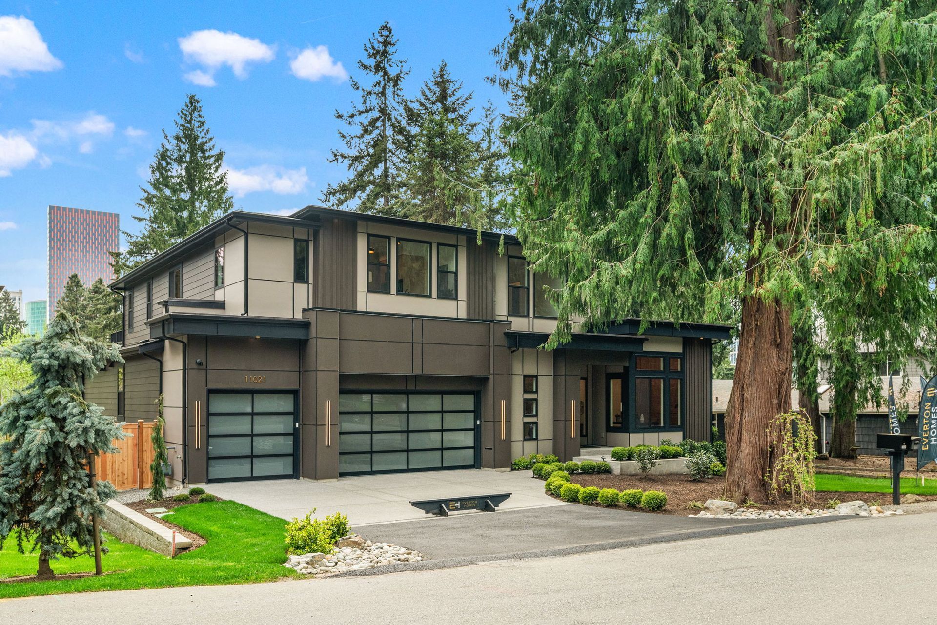 A modern, two-story dark gray house with a lit two-car garage, large windows, and a surrounding lawn at twilight.