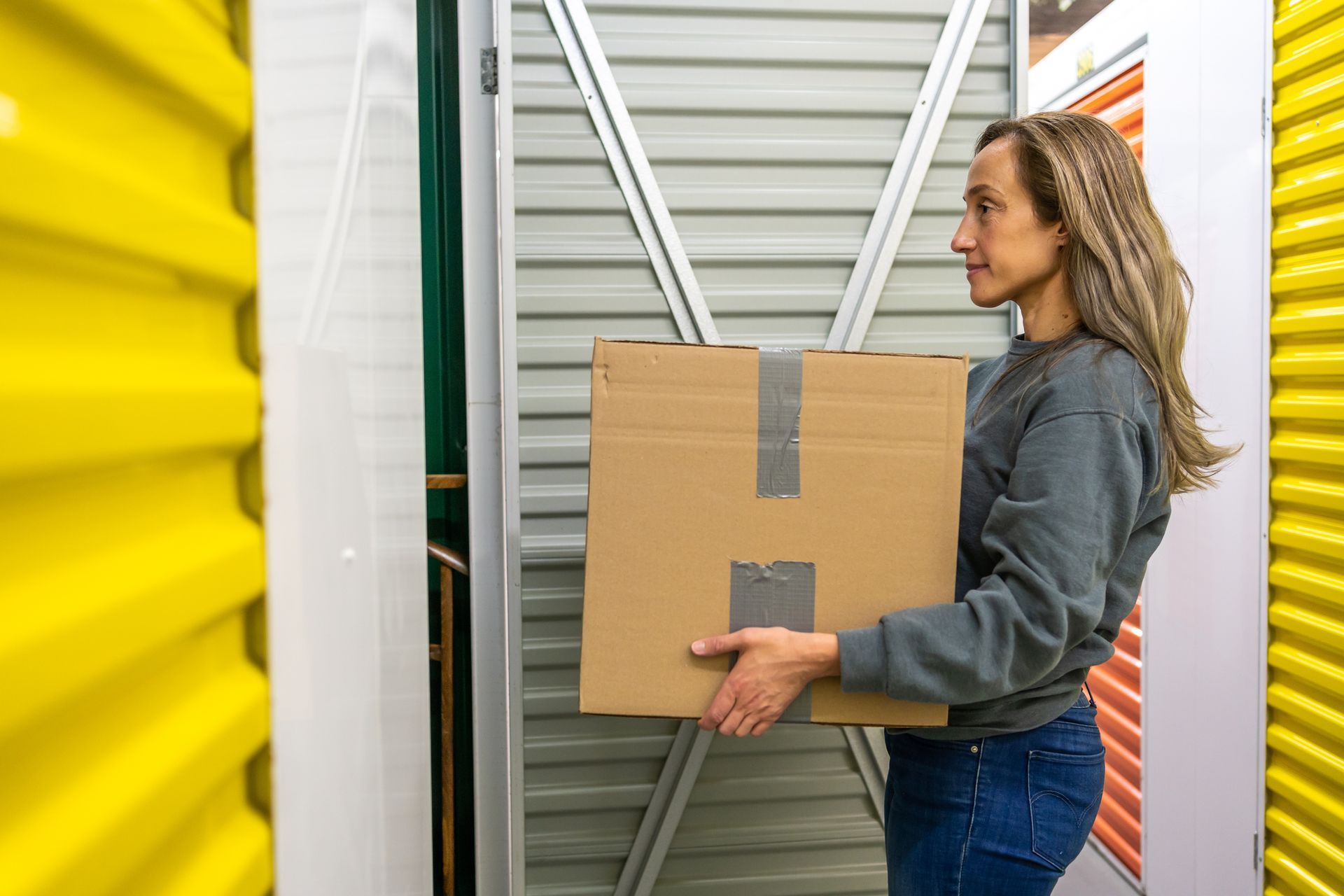 A woman in a gray sweater carries a box into her mini storage unit, with colorful doors behind.