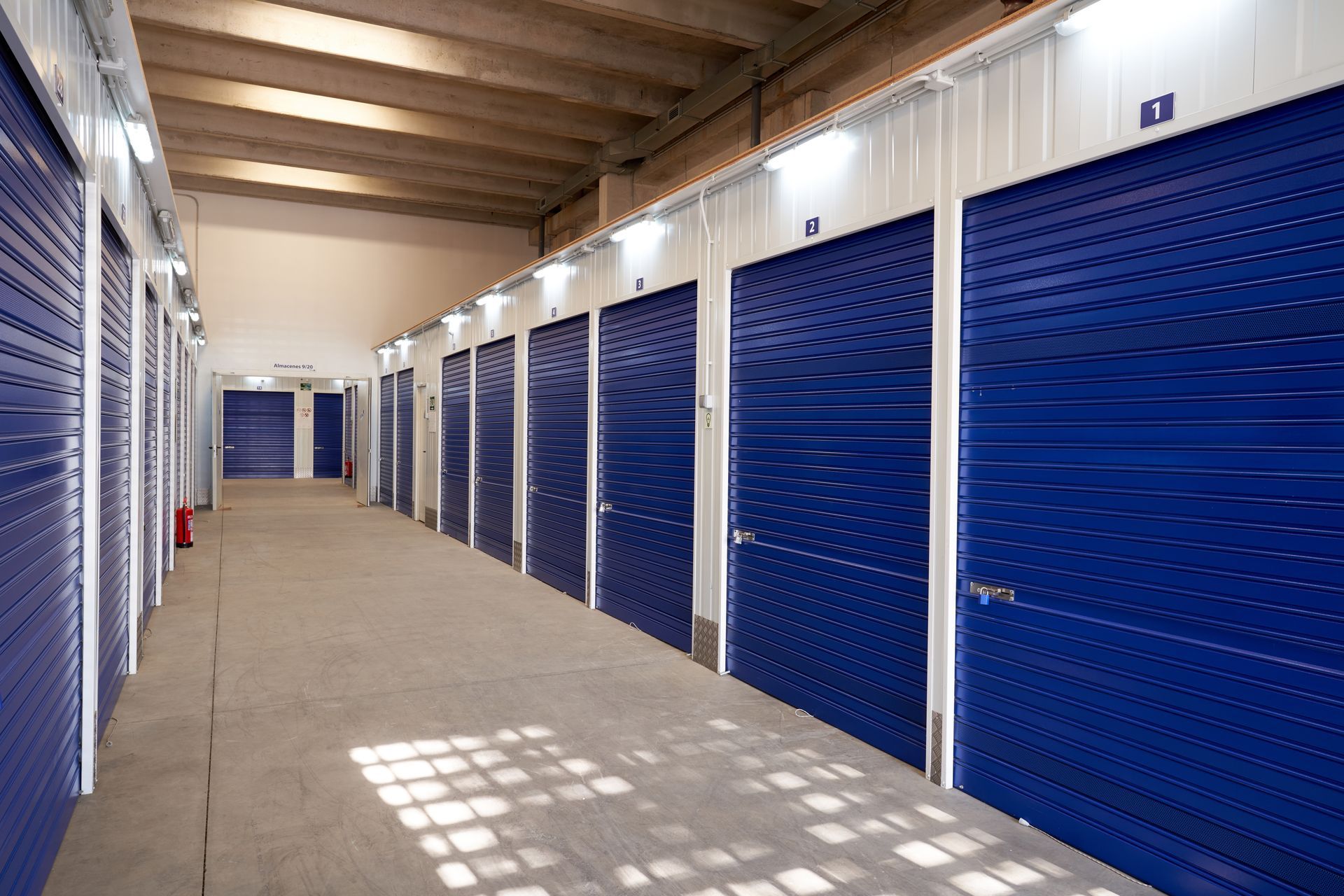 Interior hallway of numbered storage units with blue roll-up doors and lighting.