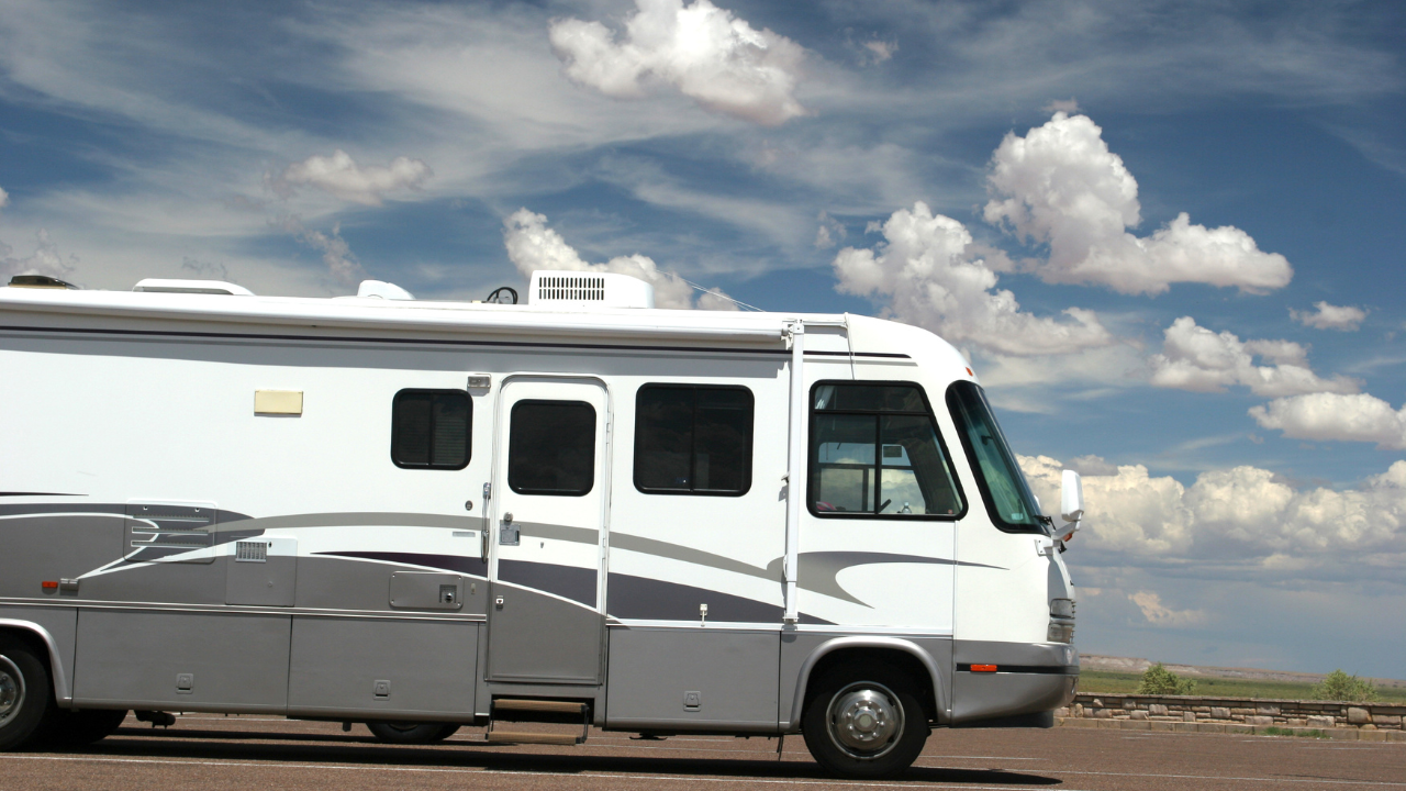 White and gray RV under a blue sky with fluffy clouds, parked on a dirt lot.