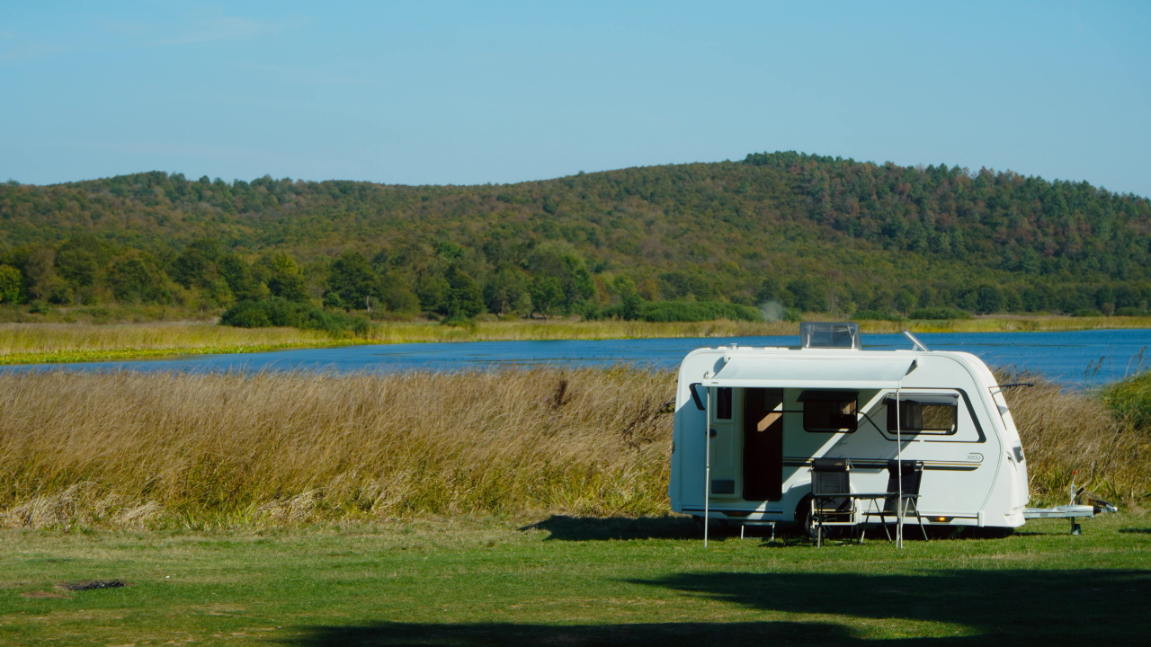 White camper trailer in a grassy field, near a lake and trees, under a blue sky.