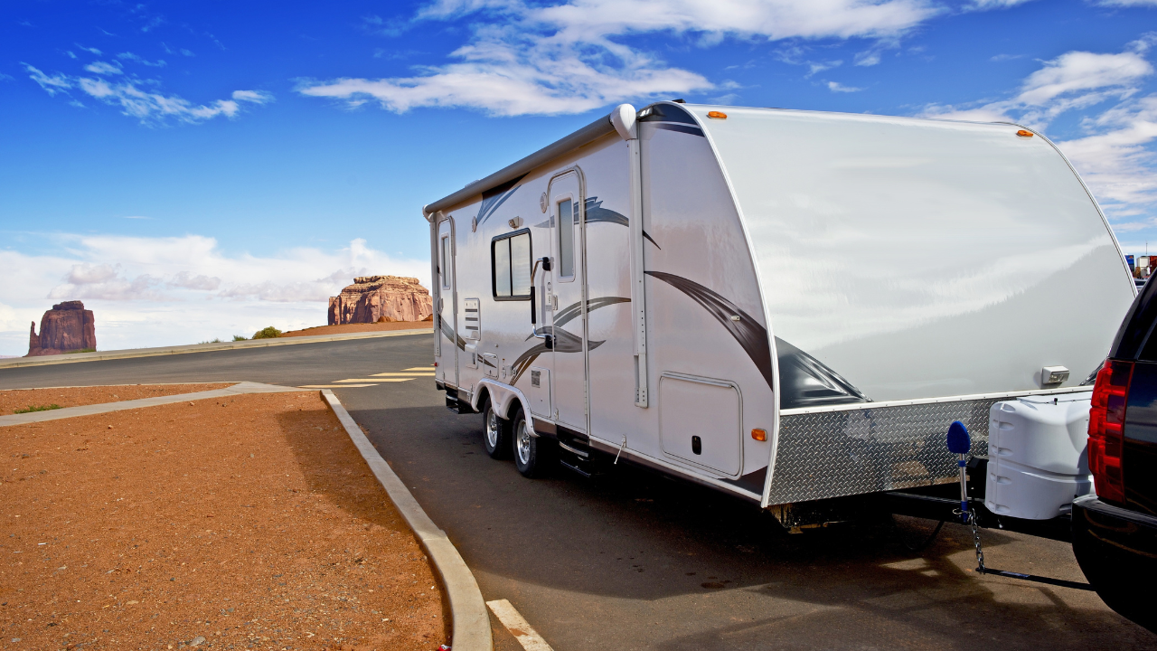 White RV hitched to a vehicle parked on asphalt, red-rock landscape, blue sky.