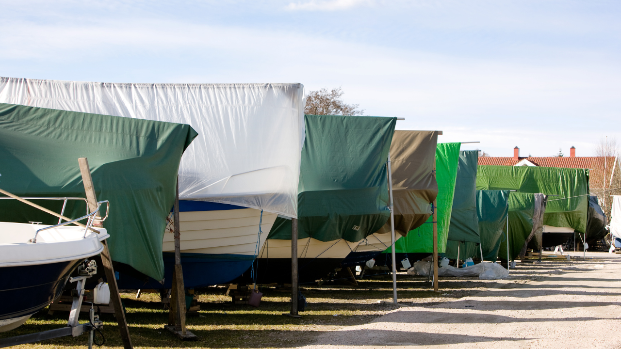 Boats covered with tarps, lined up in a row near a body of water under a bright sky.