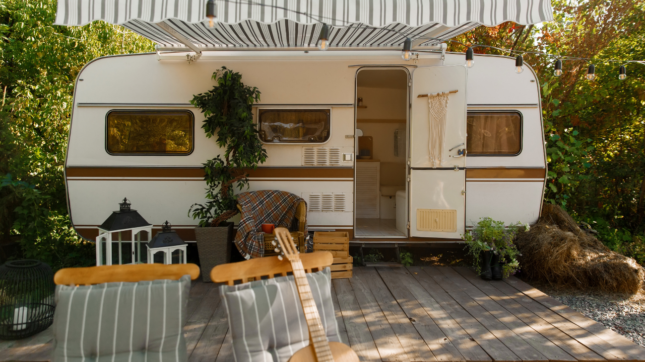 White camper with wooden deck, awning, chairs, and a guitar, set outdoors.
