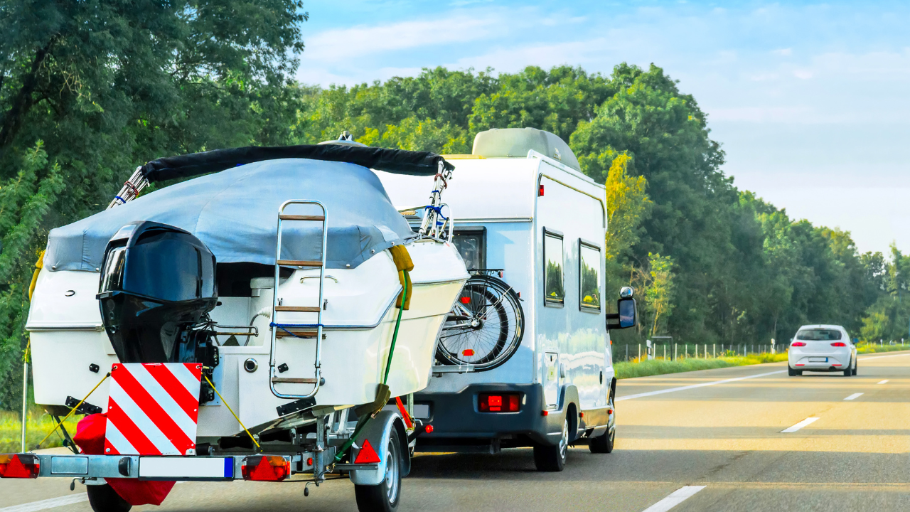 RV towing a boat on a highway, trees in the background, sunny day.