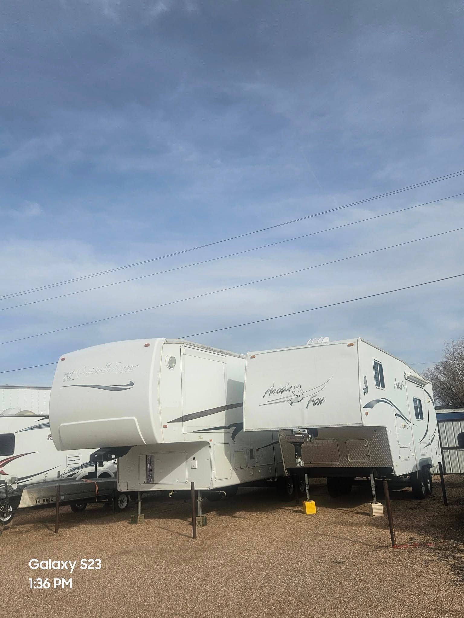 White and gray fifth-wheel RVs parked on gravel under a partly cloudy sky.