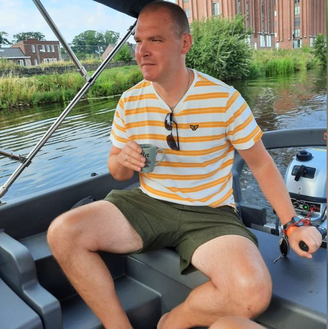 Een persoon zit in een boot, houdt een glas vast, draagt een gestreept shirt en een groene korte broek, en vaart op een rustige rivier.