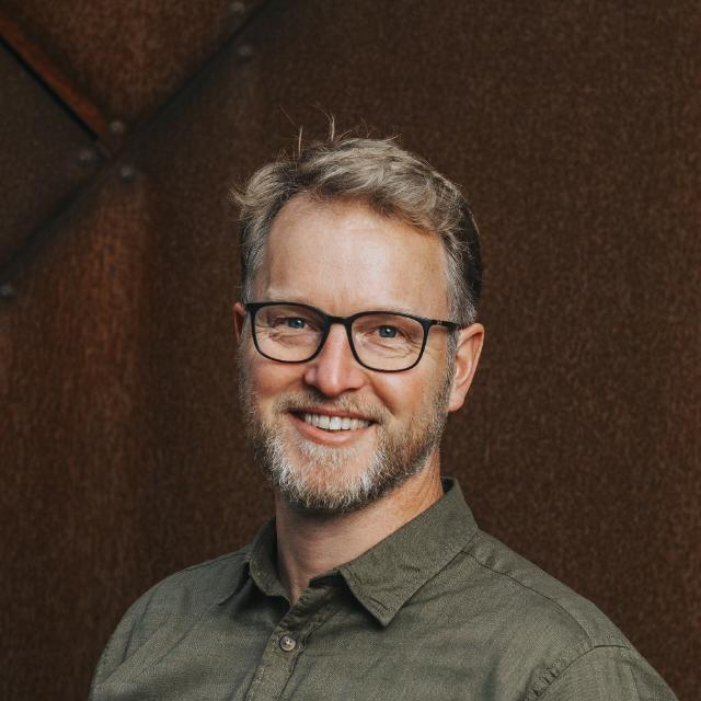 Lachende man met bril in een groen shirt tegen een donkerbruine achtergrond