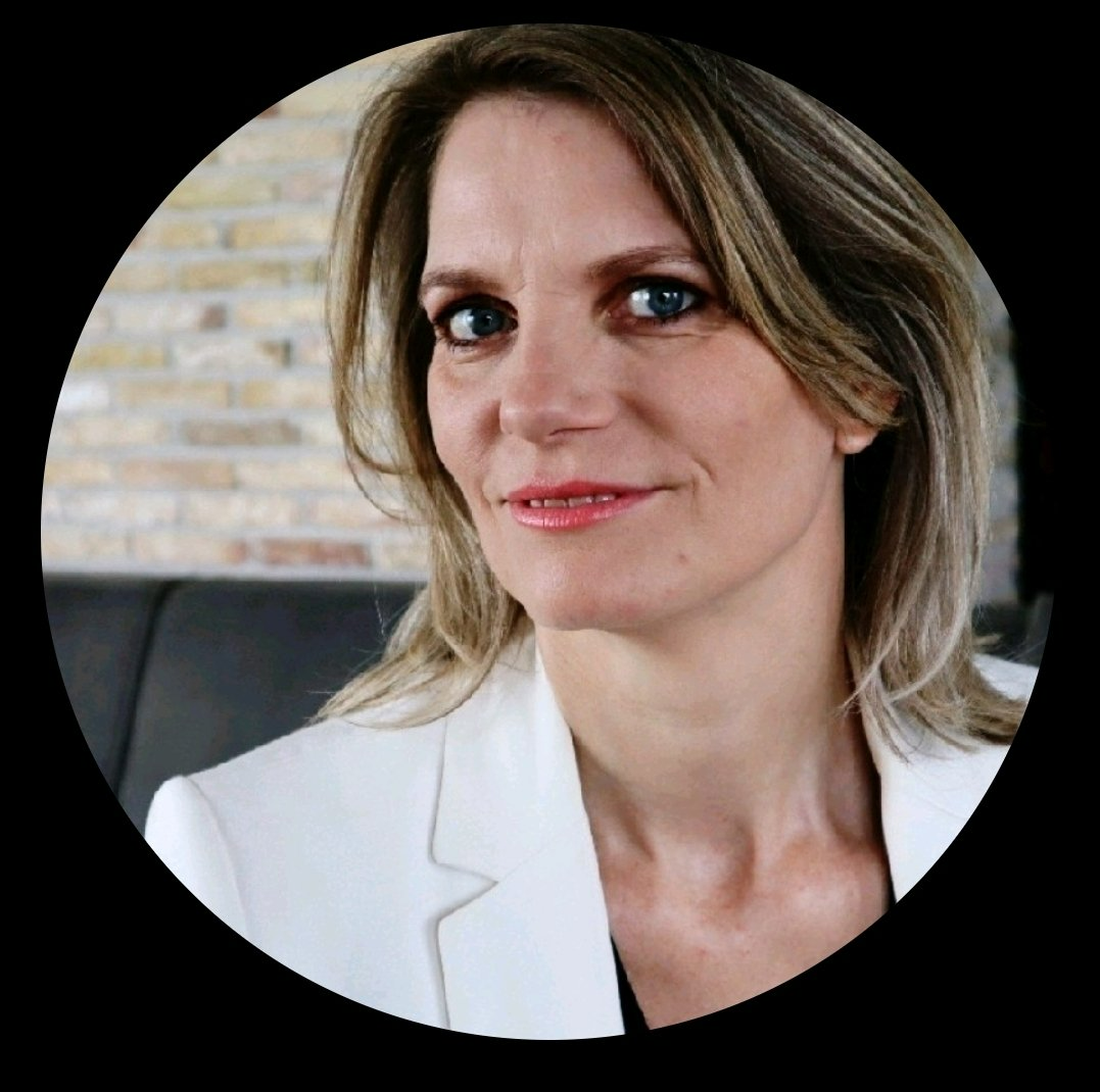 Portret van een vrouw in een witte blazer, die in de camera kijkt tegen een wazige bakstenen achtergrond.