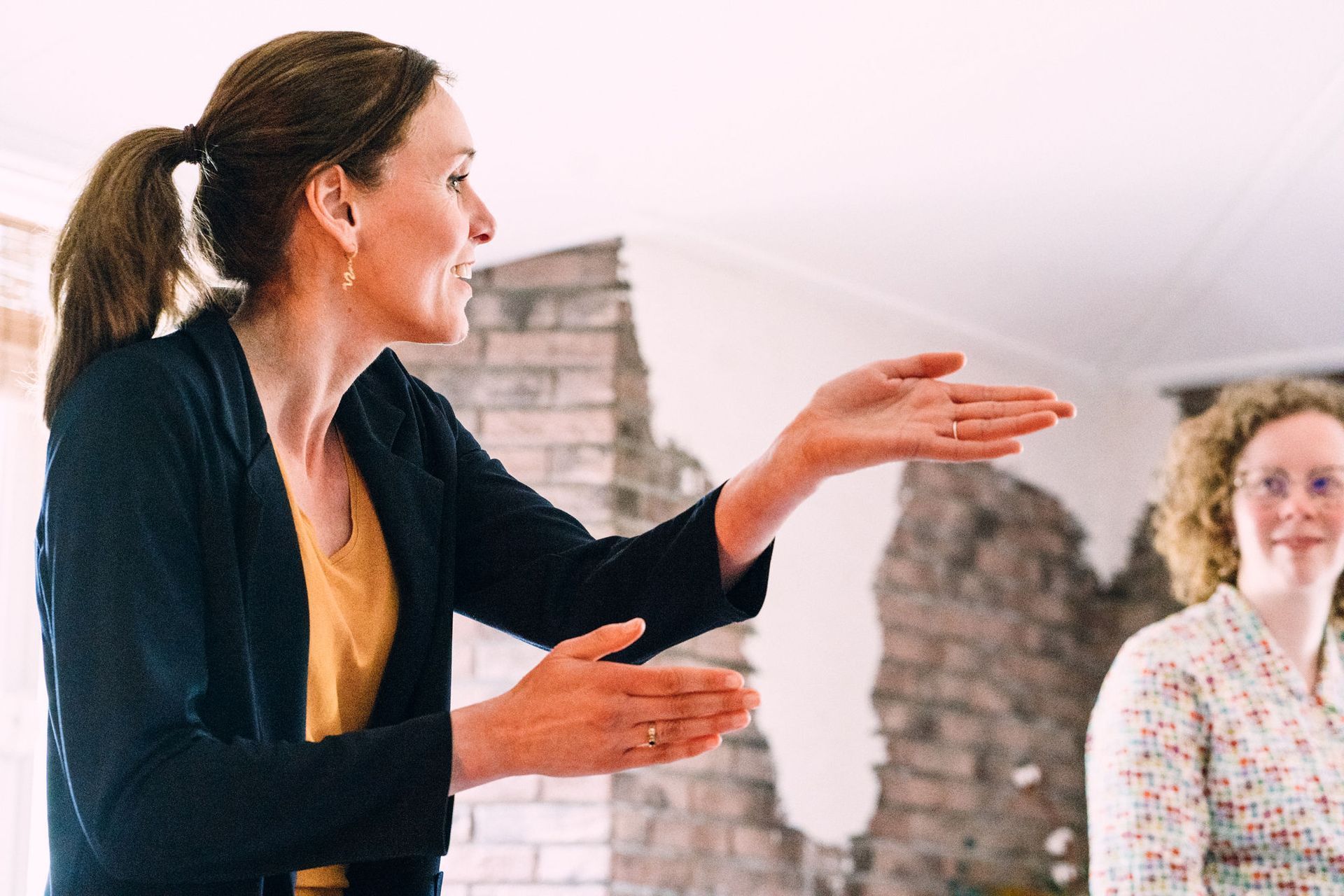 Een vrouw spreekt en gebaart in een lichte kamer, terwijl iemand anders op de achtergrond luistert.