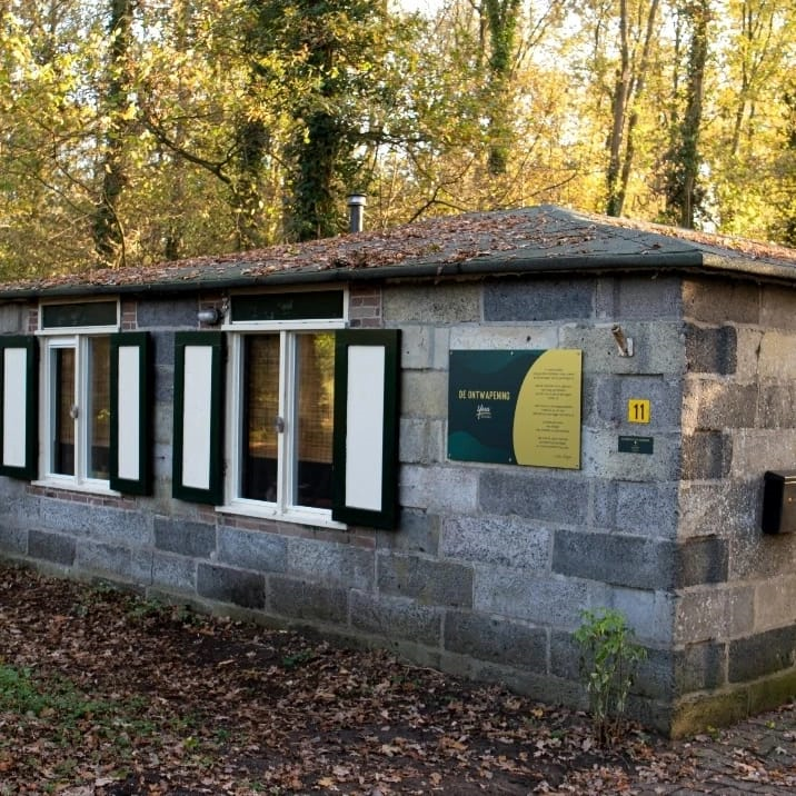 Klein stenen gebouw in een bosrijk park, met zwart-witte ramen en een groen bord aan de muur.