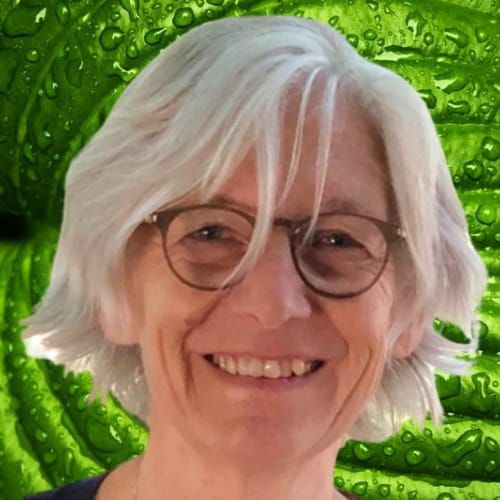 Lachende vrouw met wit haar en een bril tegen een groene, bladerrijke achtergrond.