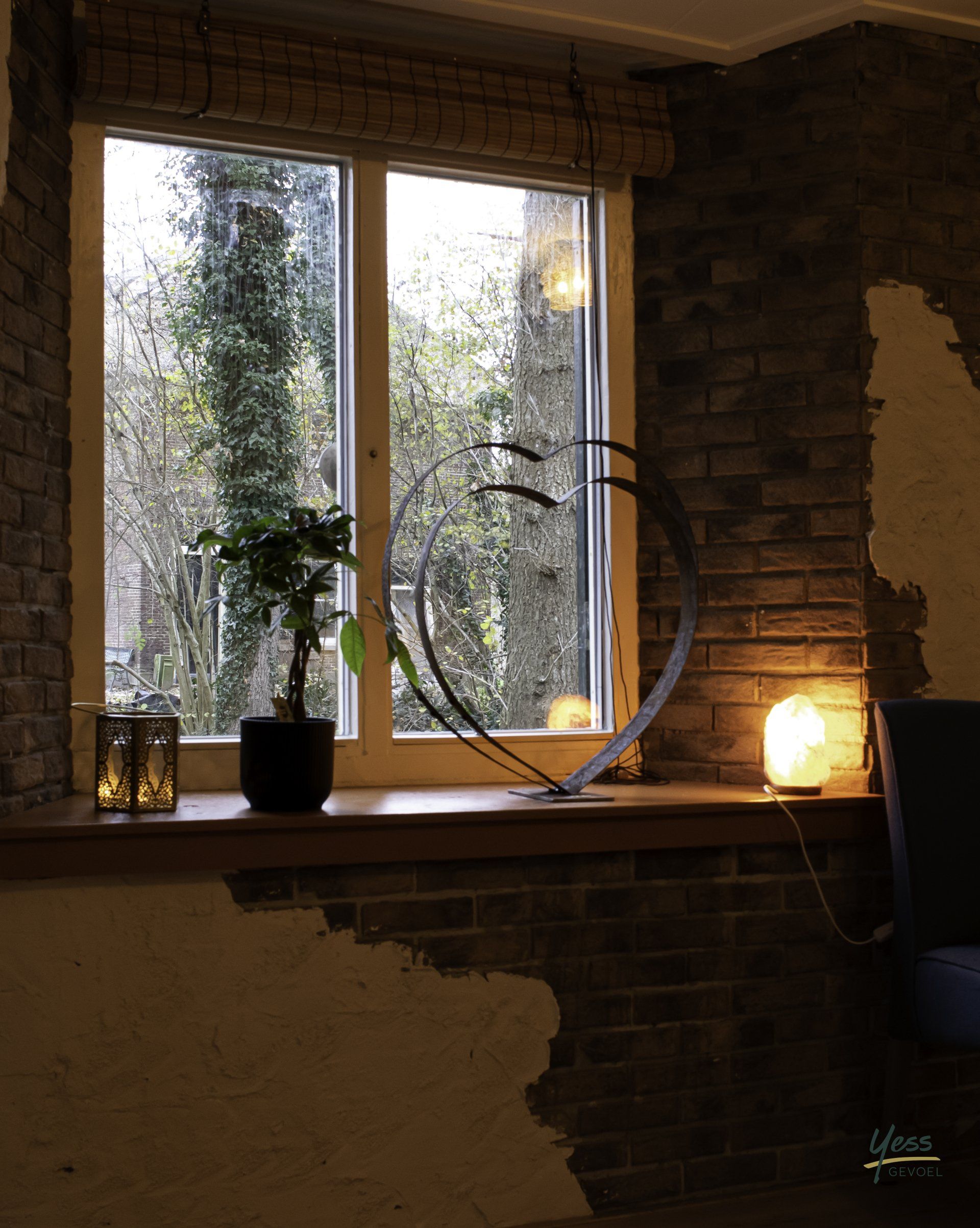 Gezellige kamerhoek met raam, potplant, rond beeldhouwerk en sfeervolle lamp op een bakstenen vensterbank.