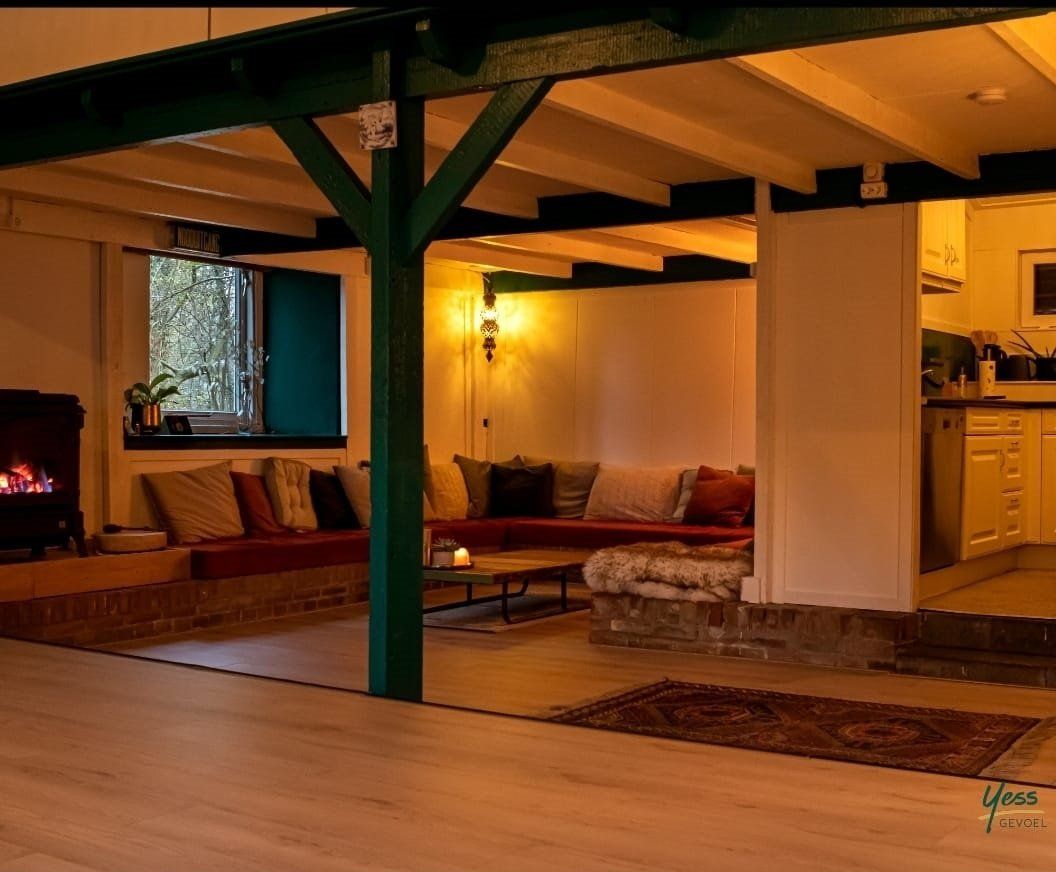 Gezellige woonkamer met warme verlichting, comfortabele banken, een open haard en groene draagbalken.
