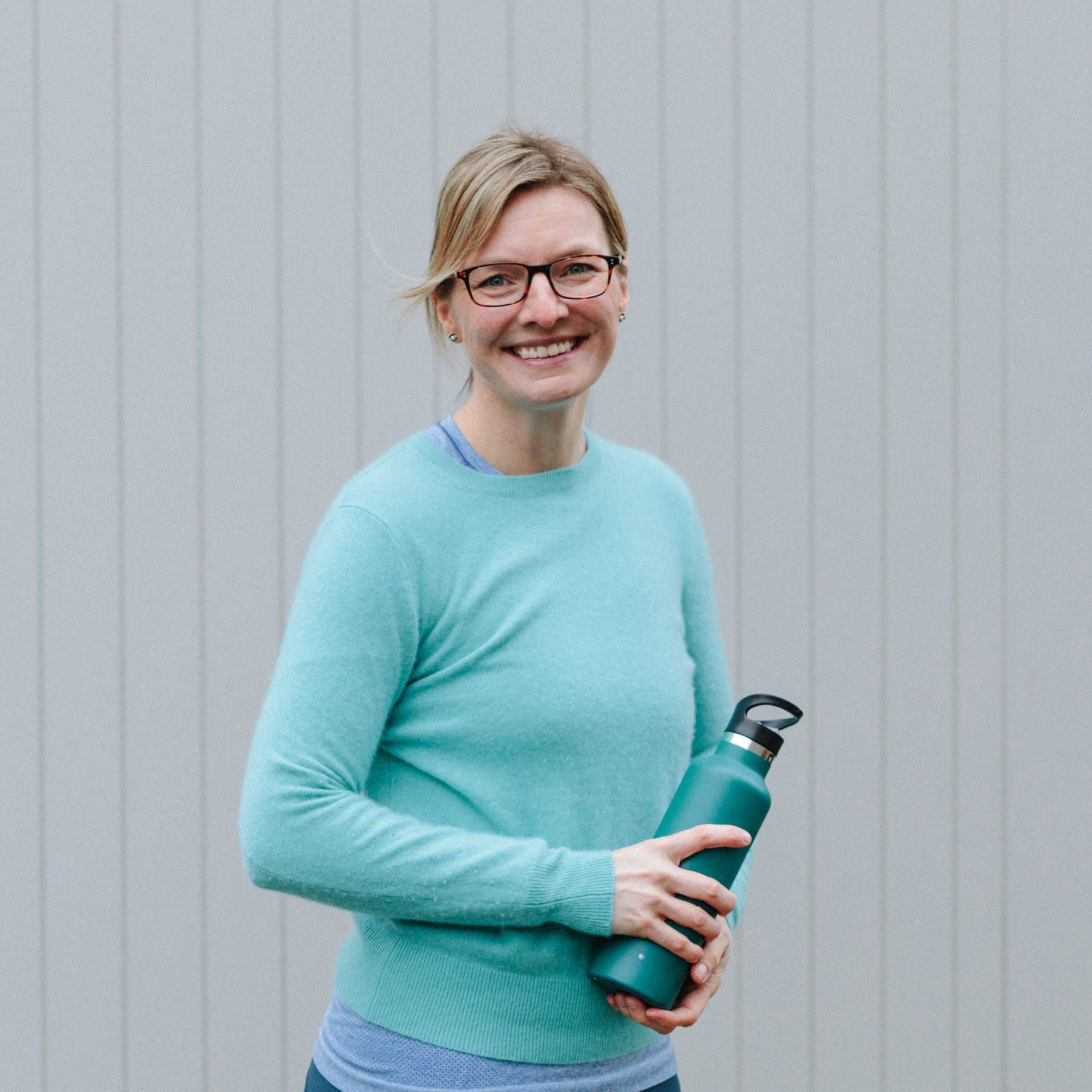A woman wearing glasses and a blue sweater is holding a green water bottle.