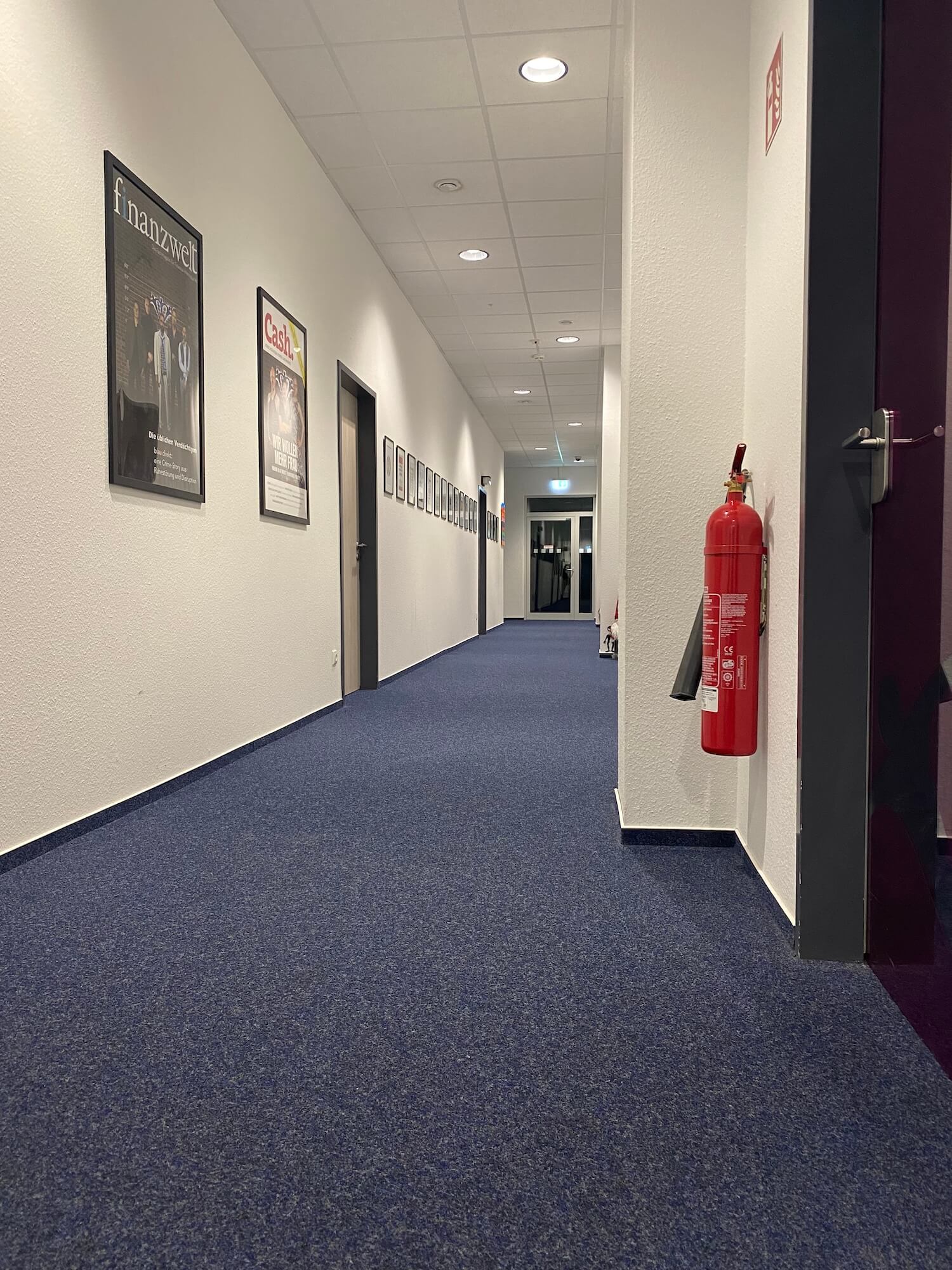 Ein langer Flur mit blauem Teppich und einem roten Feuerlöscher an der Wand.