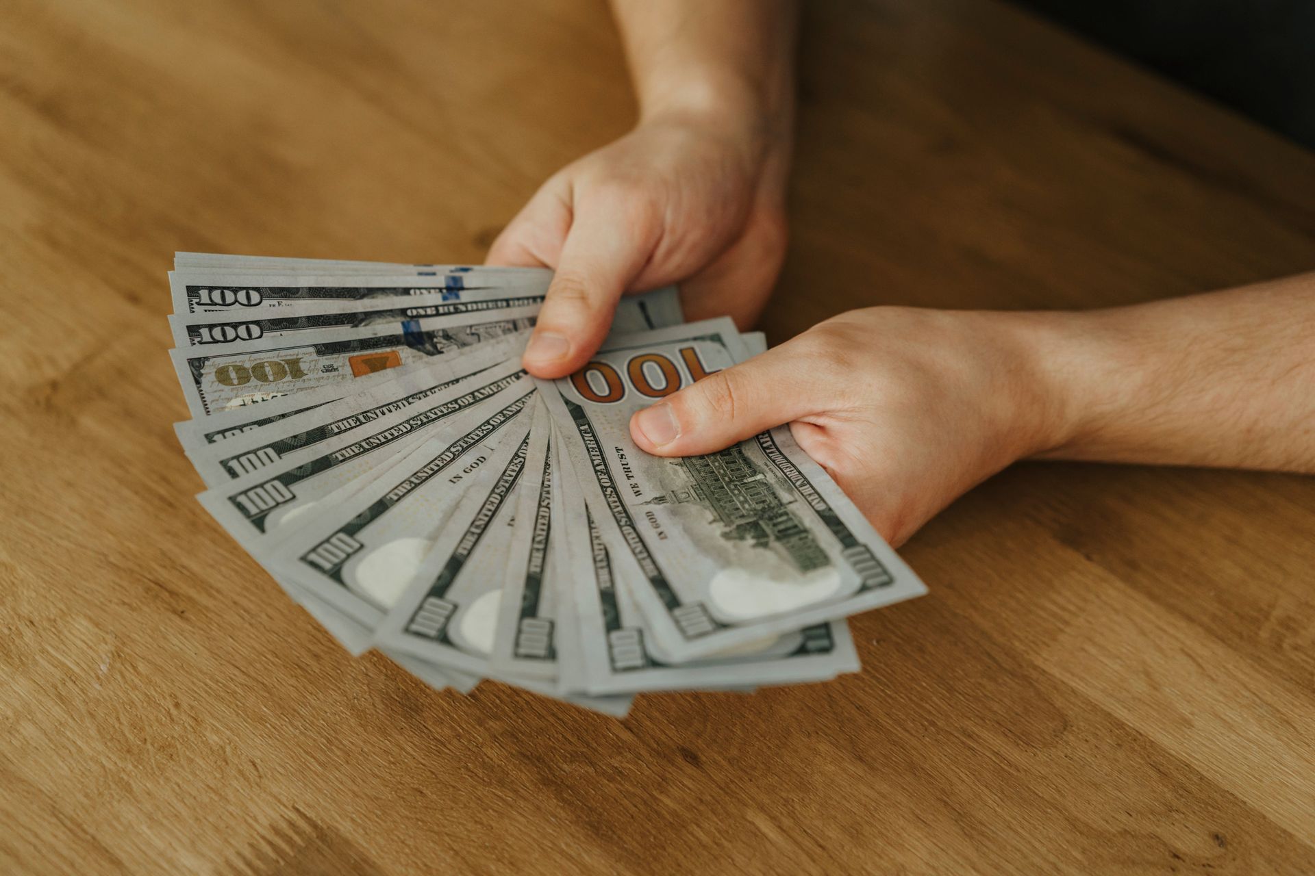 Hands fanning several U.S. hundred-dollar bills over a wooden table
