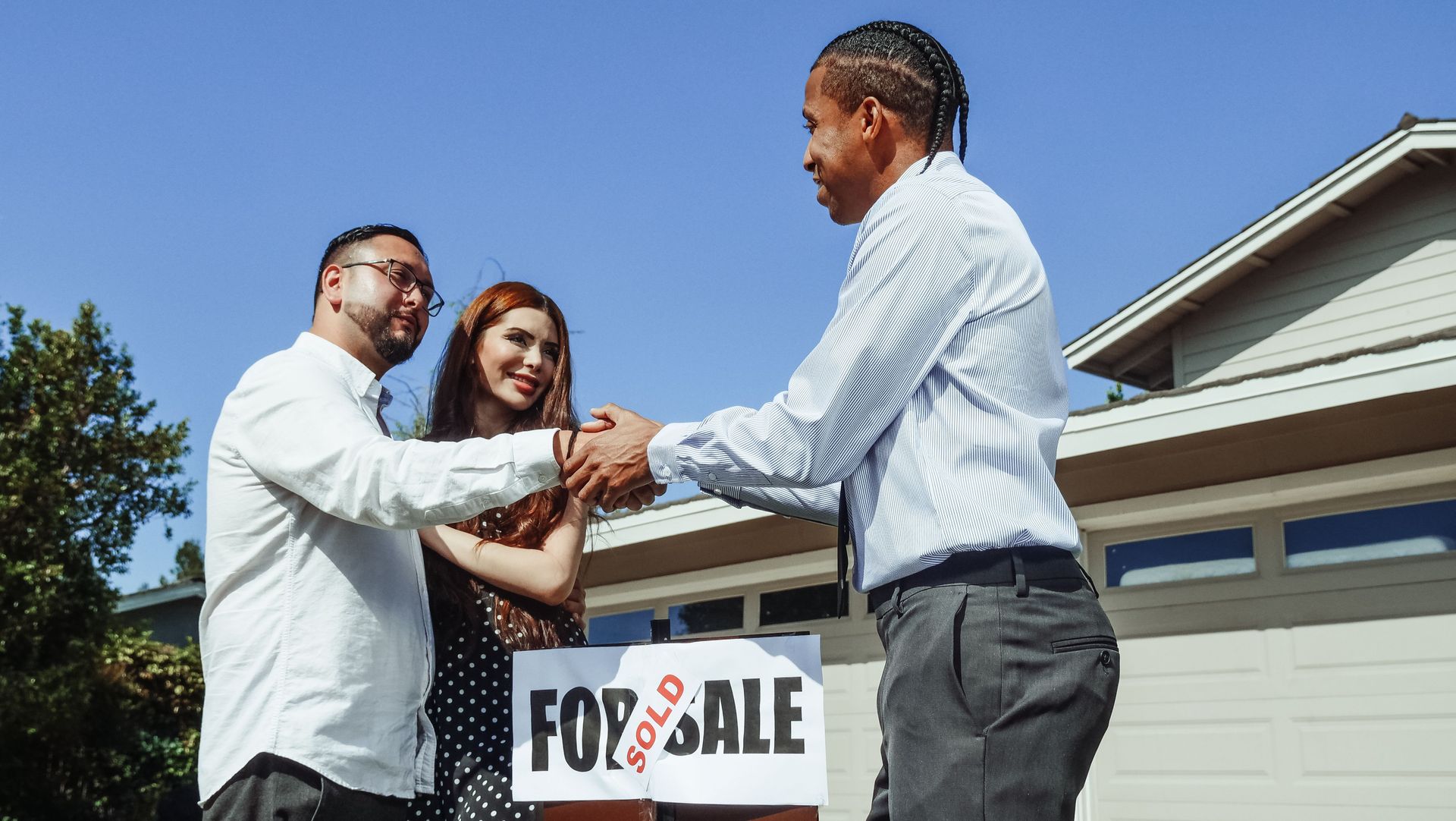 Two people shaking hands with a real estate agent beside a “For Sale” sign in front of a house