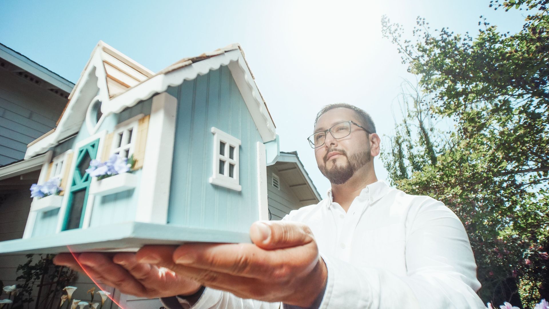 Man holding a small blue house model outdoors in bright sunlight
