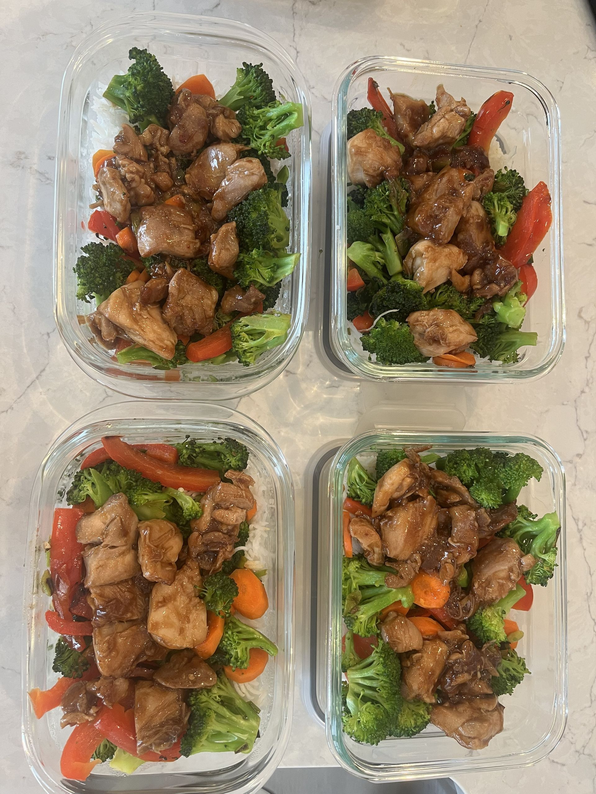 Four meal prep containers with cooked meat, broccoli, carrots, and greens arranged on a white surface