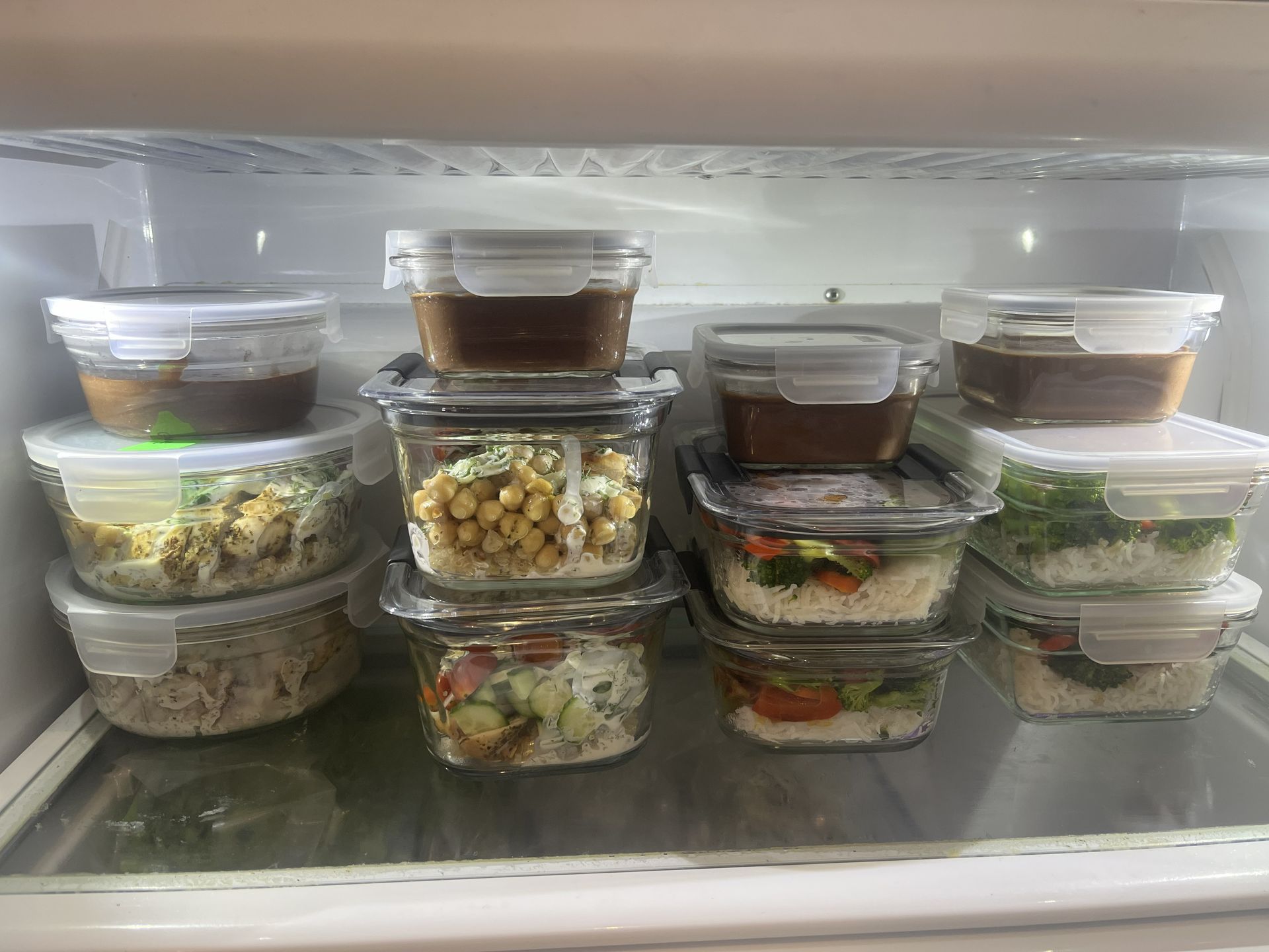 Stacked clear meal prep containers in a refrigerator, holding assorted salads and leftovers.