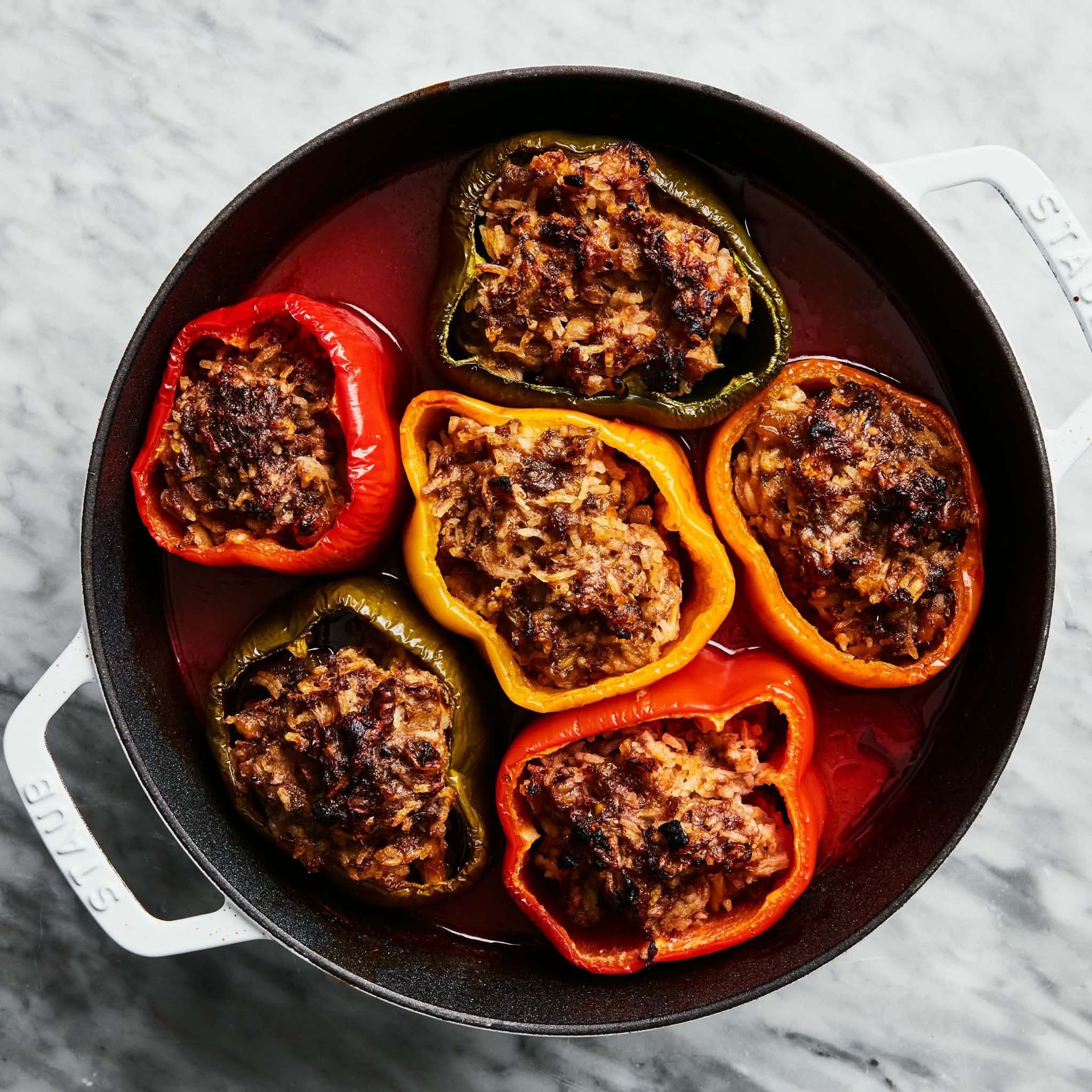 Stuffed bell peppers in a cast-iron skillet, with red, yellow, and green peppers on a marble surface
