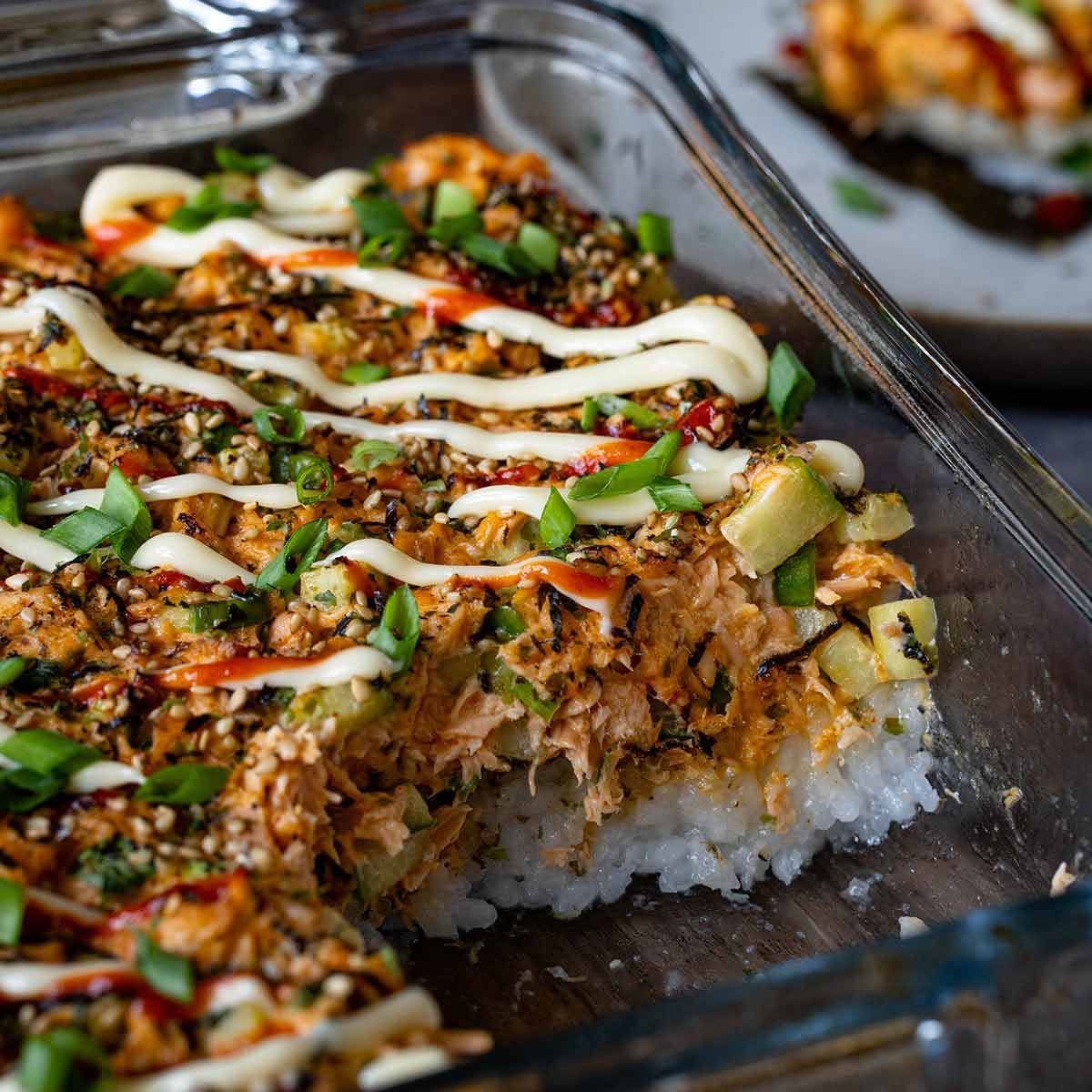A glass dish of sushi rice topped with shredded meat, chopped herbs, and drizzled sauces