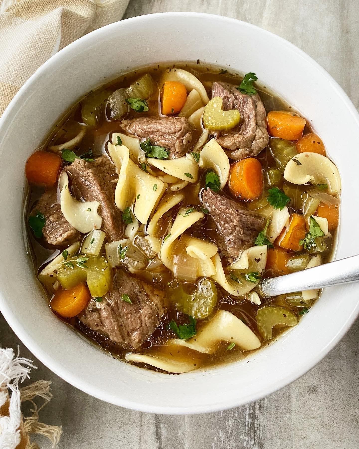 Bowl of beef noodle soup with carrots, celery, and herbs on a light tabletop