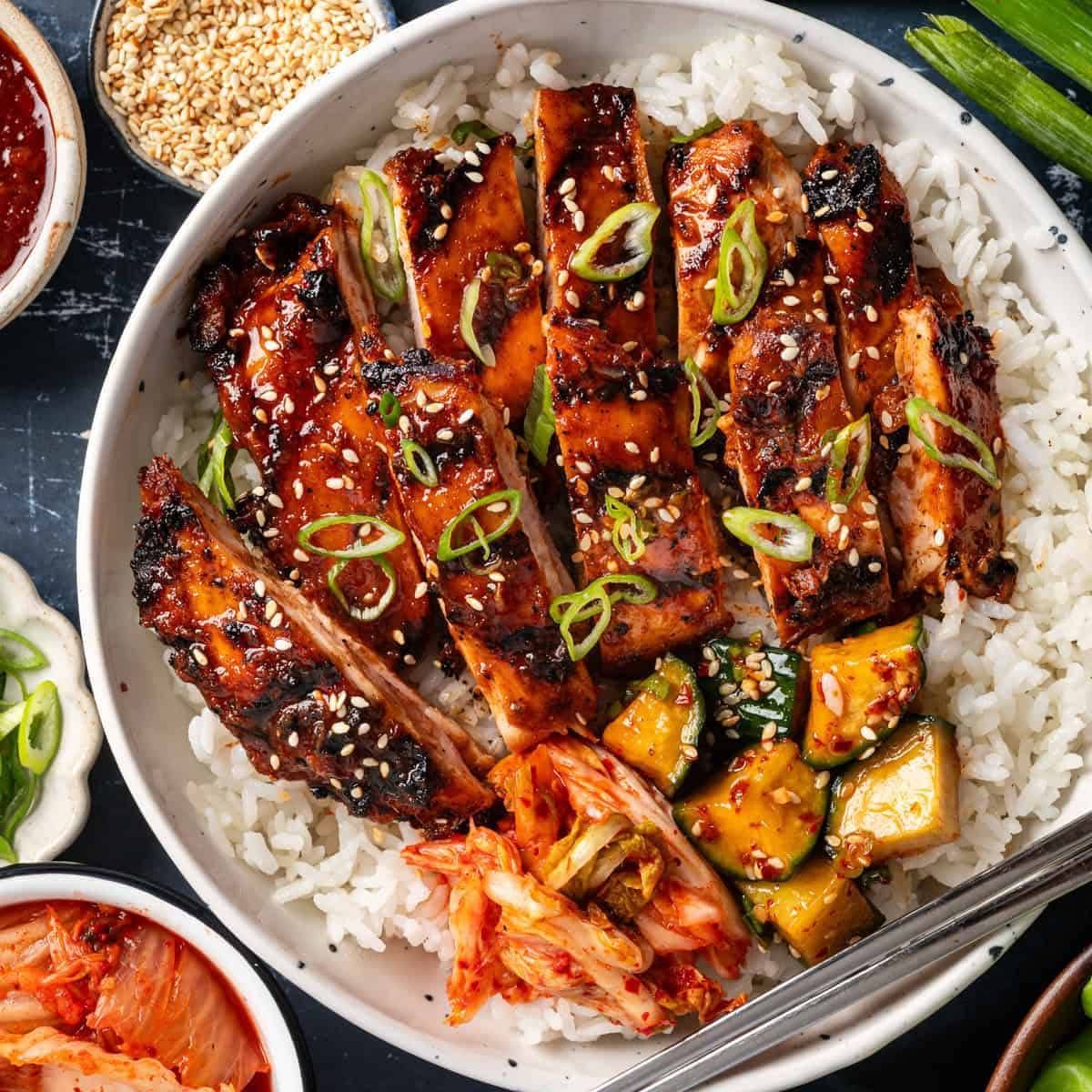 Rice bowl with glazed grilled chicken, kimchi, zucchini, and green onions, with side dishes and chopsticks