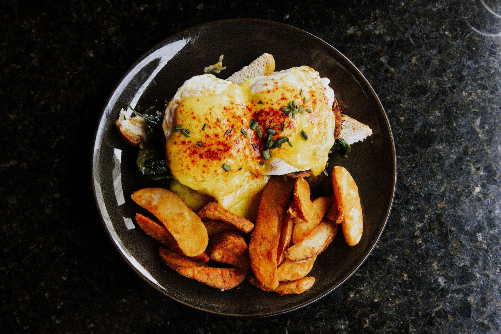 A serving of Eggs Benedict with hollandaise sauce and potato wedges on a dark plate against a black granite background.