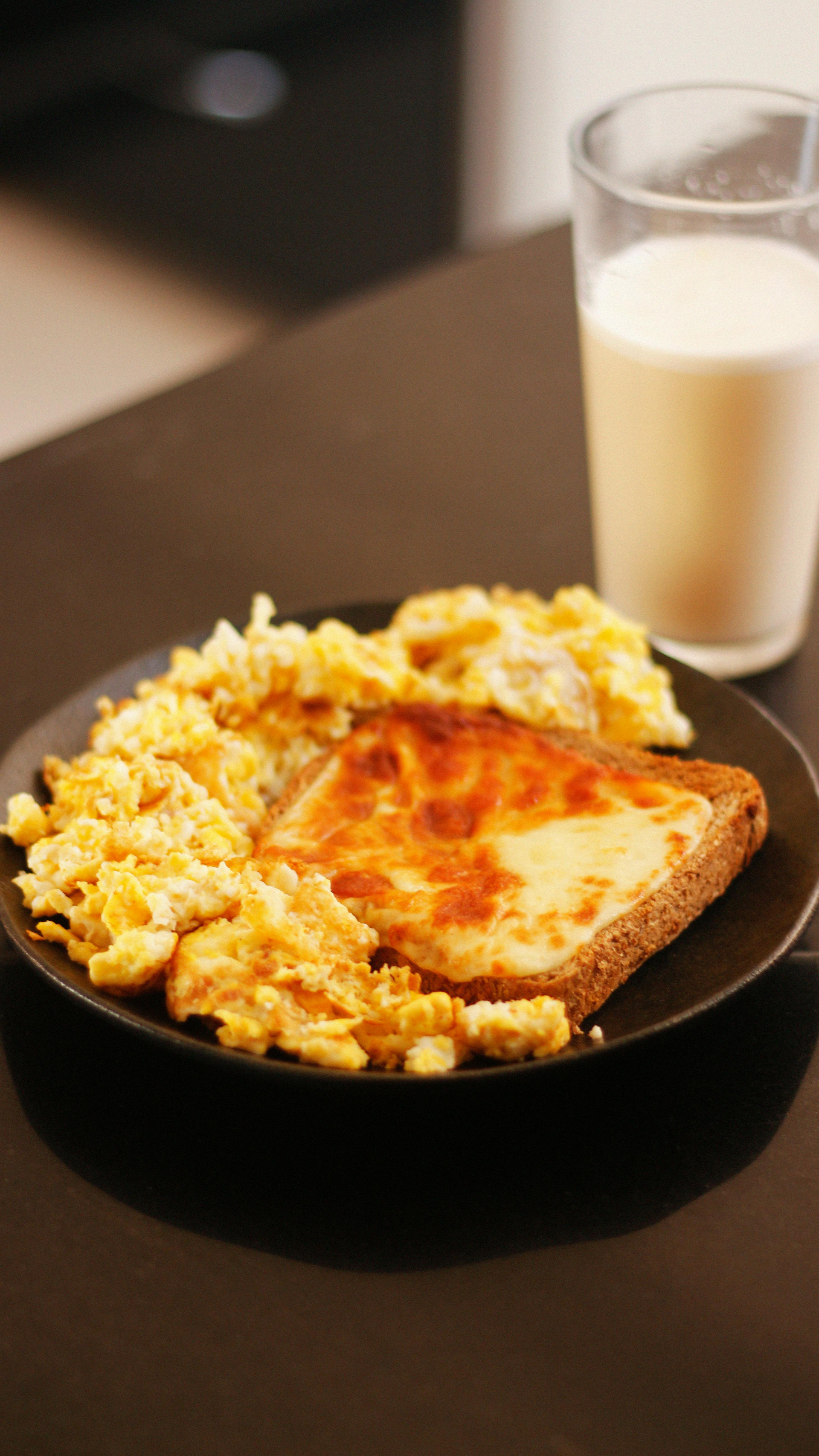 A plate of scrambled eggs with a piece of cheesy toast next to a glass of milk on a dark table.