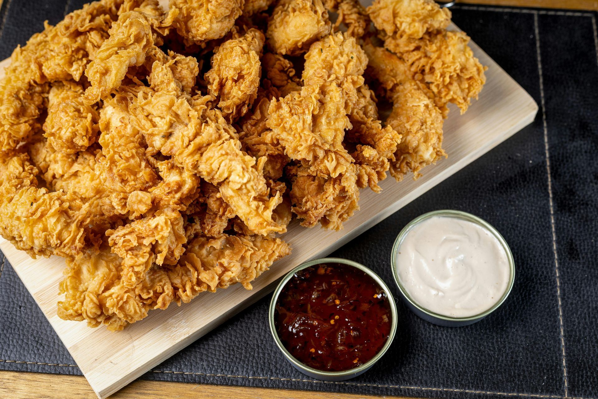 A wooden board piled with golden-brown fried chicken strips, served with small bowls of dark red and white dipping sauces.