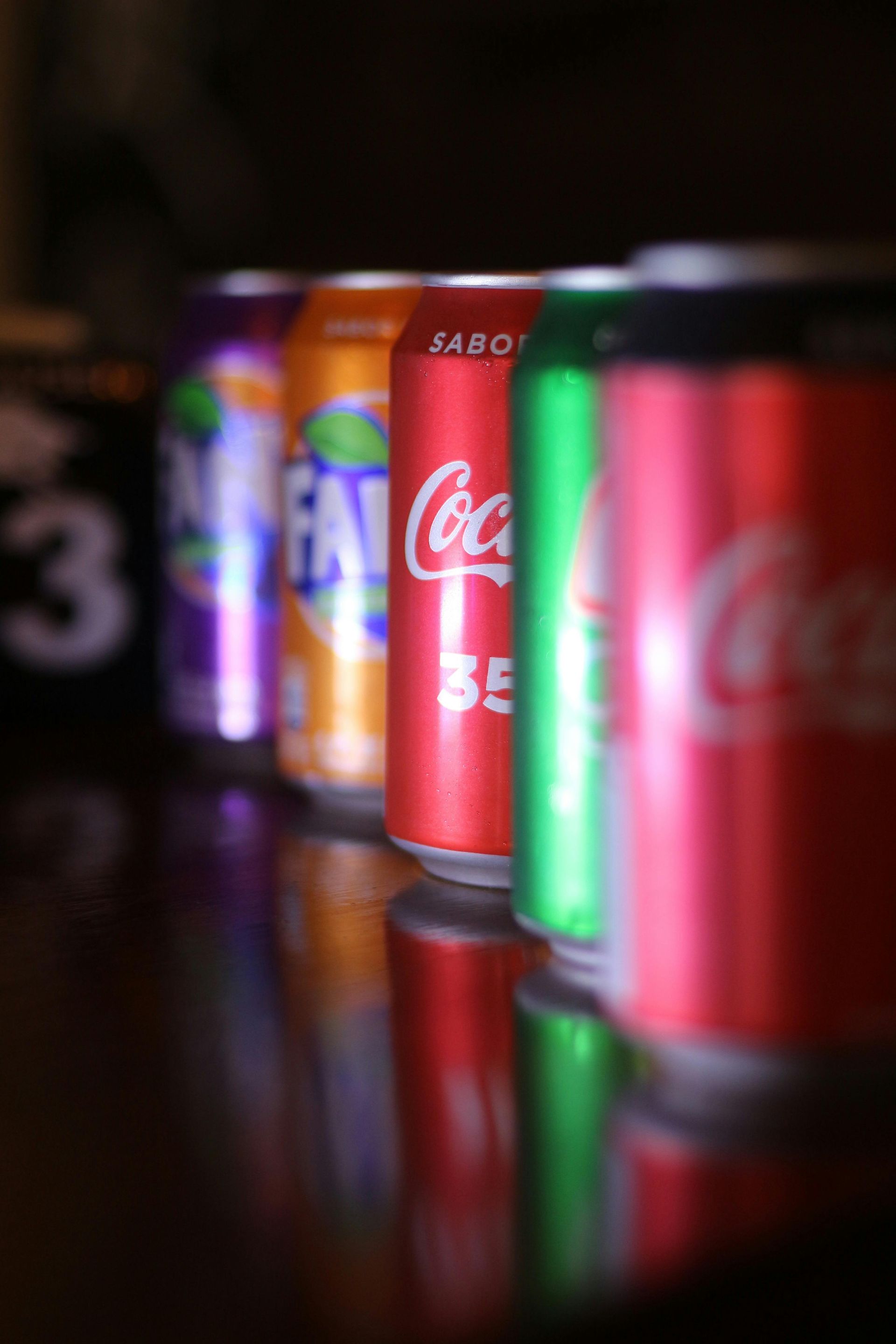 A row of colorful soda cans stands on a reflective surface, with focus on a red Coca-Cola can in the middle.