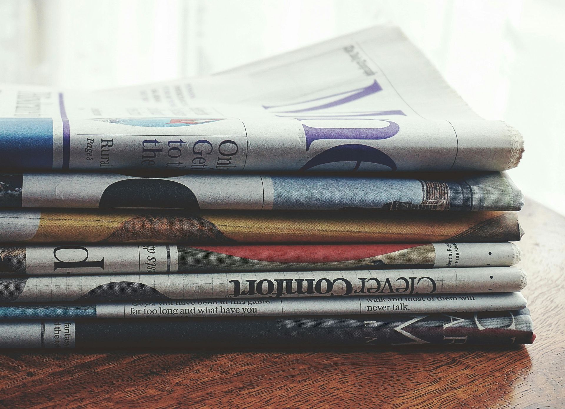 Pile of folded newspapers on a wooden surface, showing various colorful mastheads and text.