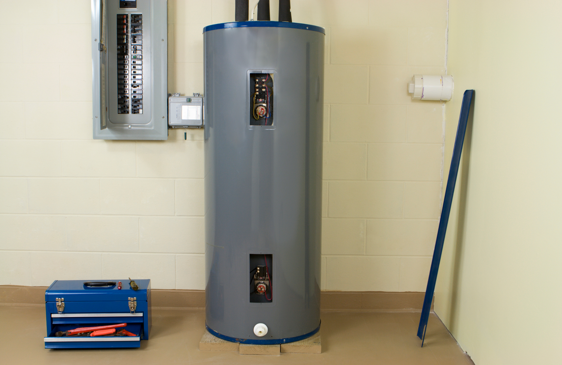 Gray water heater in a basement next to an electrical panel and blue toolbox.