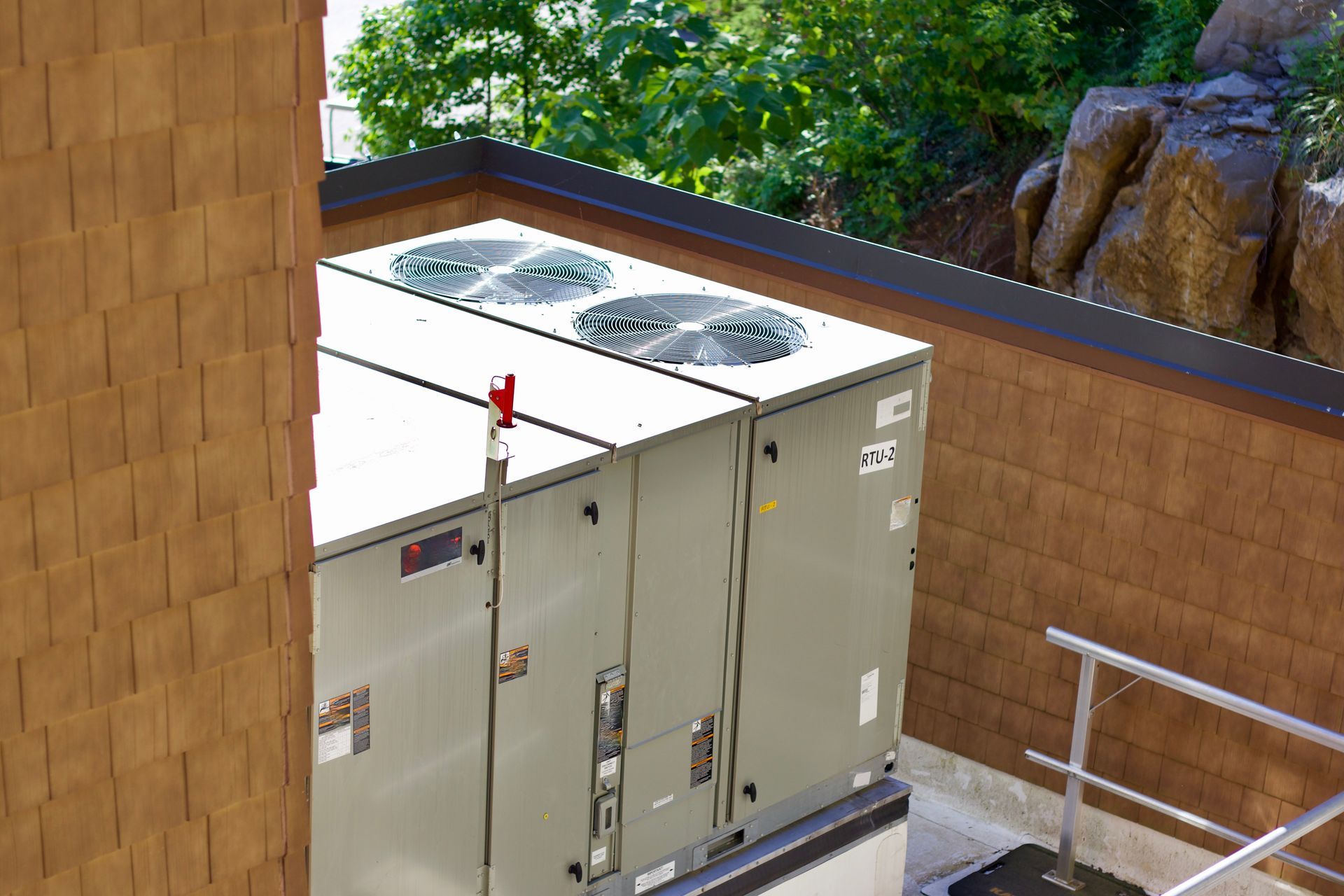Two air conditioning units on a rooftop near wooden siding and rocky terrain.