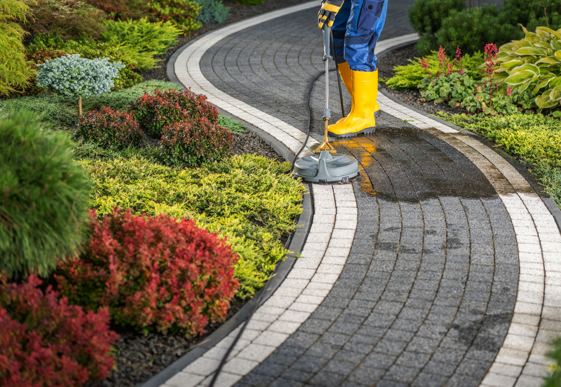 A man in yellow boots is cleaning a brick walkway in a garden.