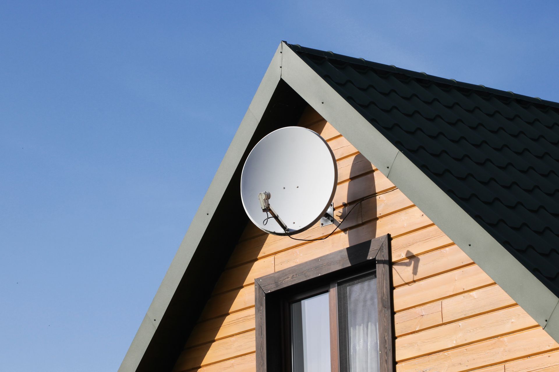 Satellite dish mounted on a wooden house's gable under a blue sky.