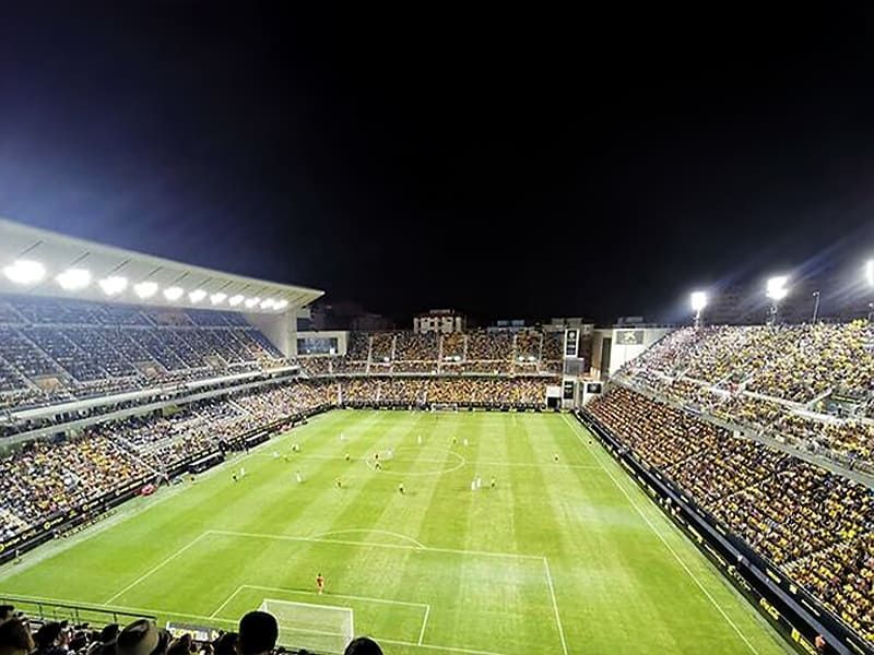 Packed soccer stadium at night with players on a bright green field and crowded stands