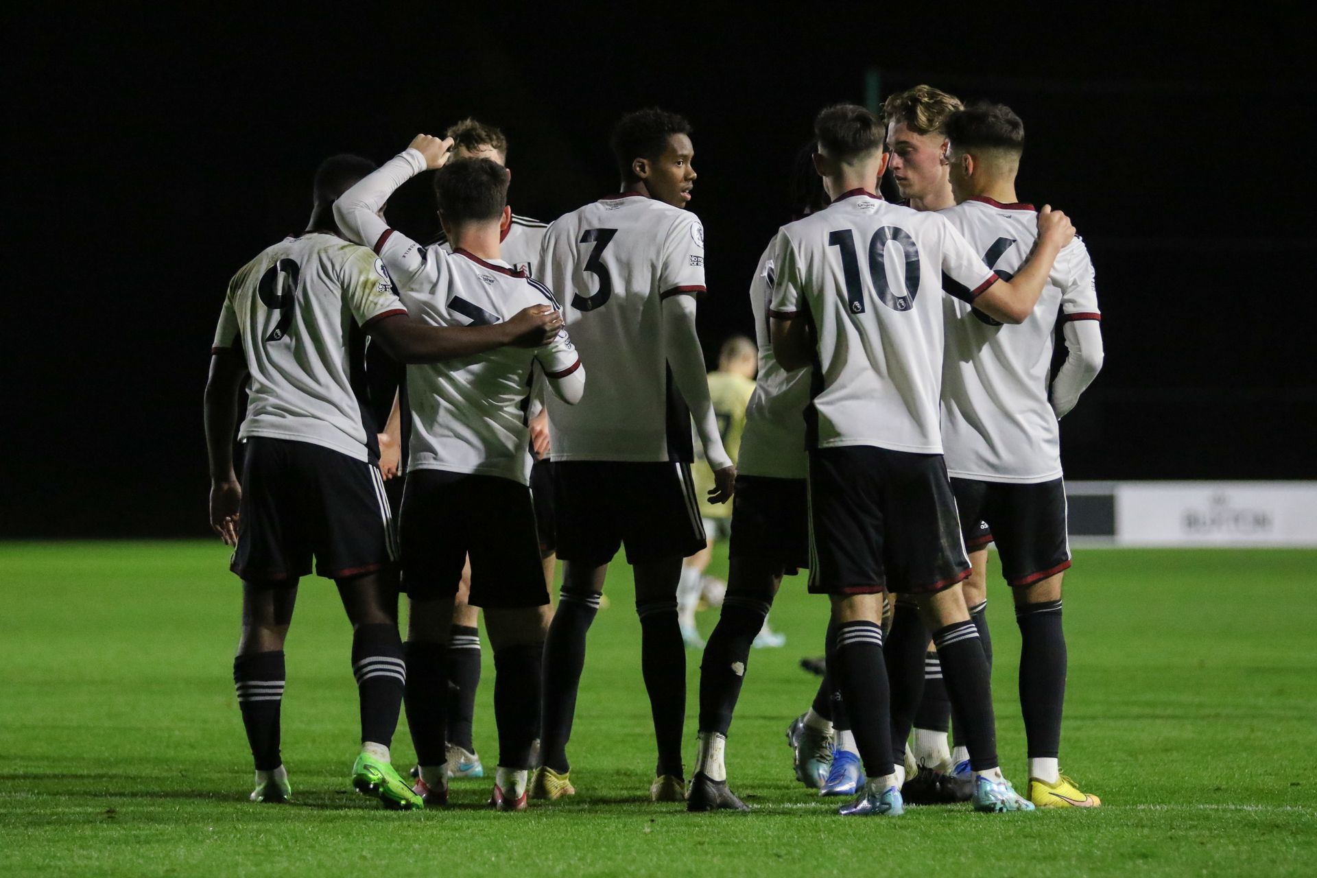 Soccer players in white jerseys huddle on a lit field at night.