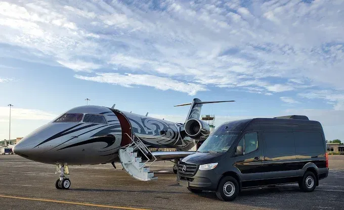 A private jet with a black van parked on the tarmac under a partly cloudy sky.