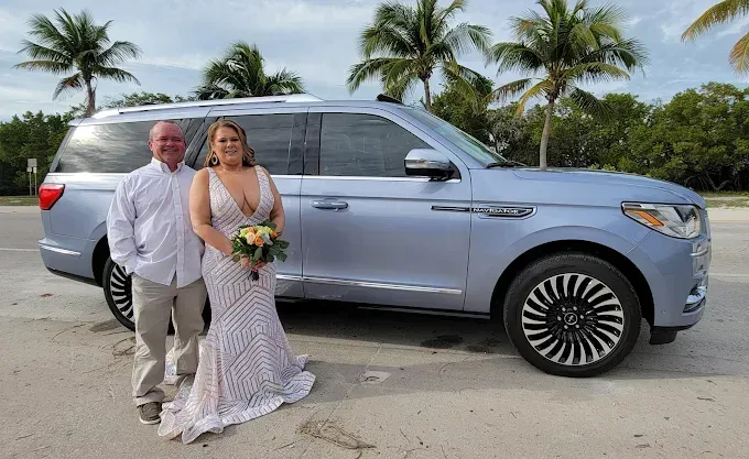 Newlyweds pose beside a light blue Lincoln Navigator. Beach setting, couple smiling.