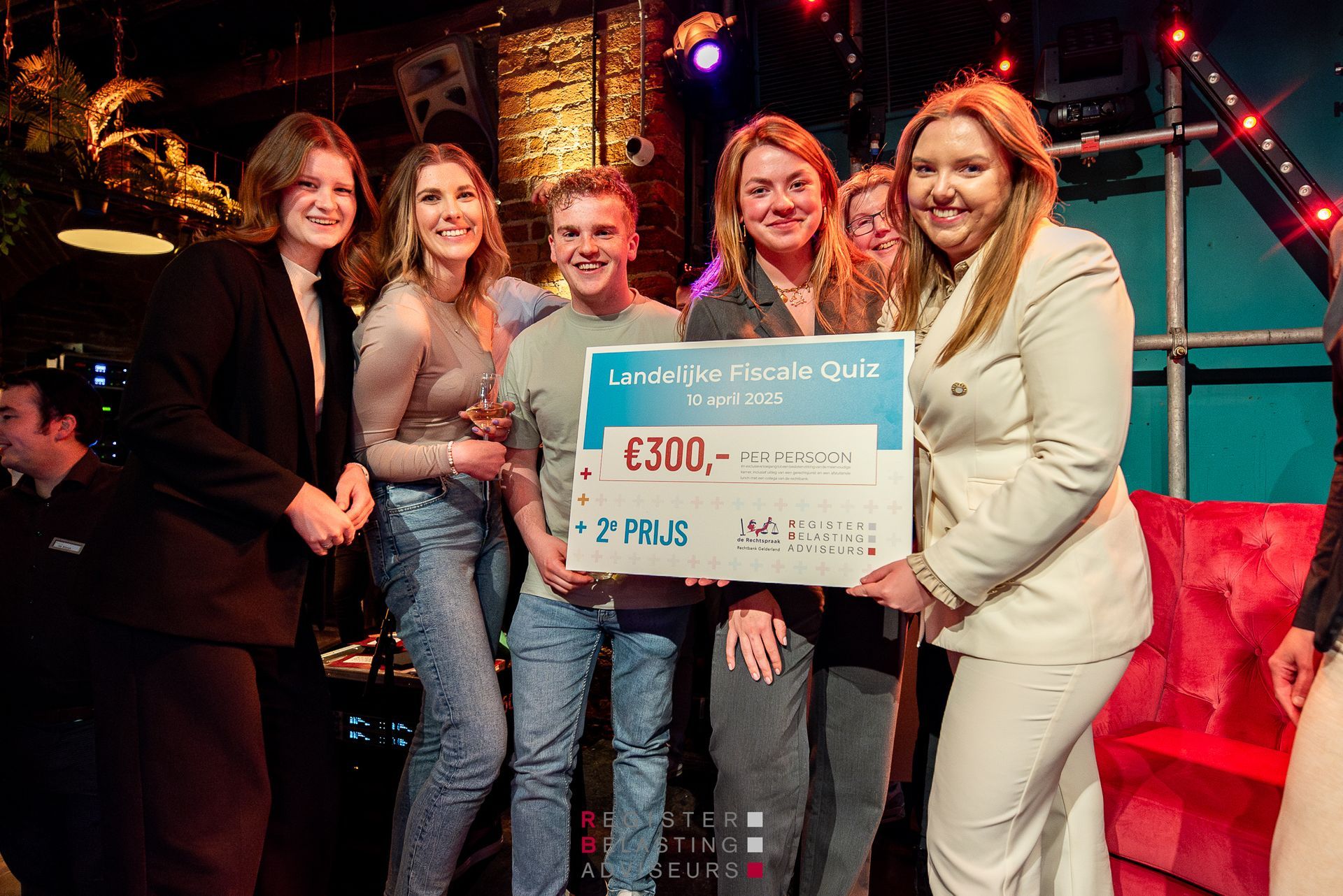 Vijf mensen glimlachen en houden een grote cheque met prijzengeld vast op een podium tijdens een evenement.