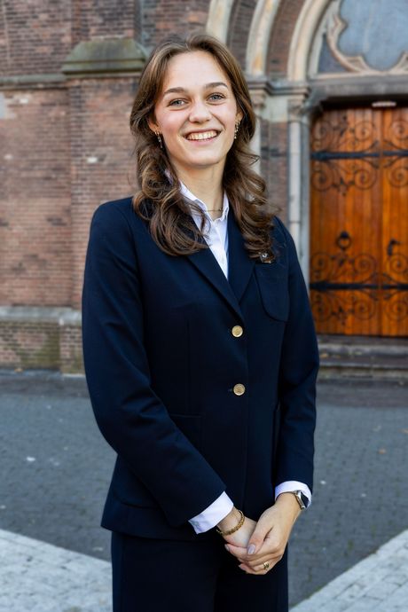 Een man in een donkerblauw pak en wit overhemd glimlacht terwijl hij voor een bakstenen gebouw staat.