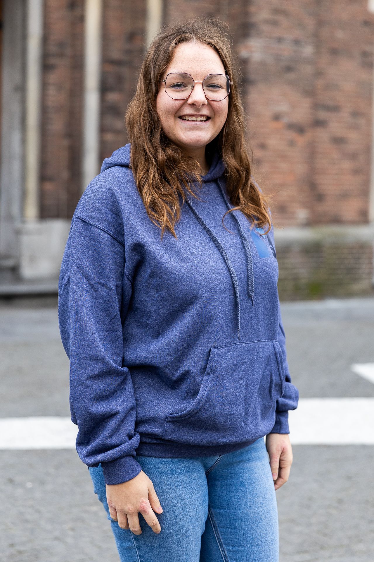 Een vrouw met een bril, een blauwe hoodie en een spijkerbroek lacht voor een bakstenen gebouw.