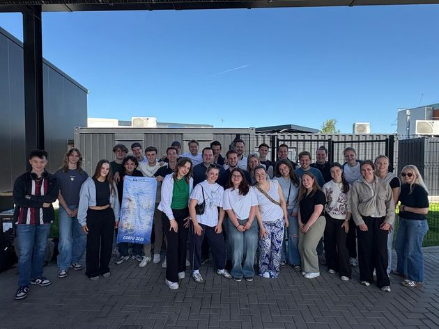Een groep mensen poseert buiten op een betegelde ondergrond, met uitzicht op een wateroppervlak onder een bewolkte hemel.